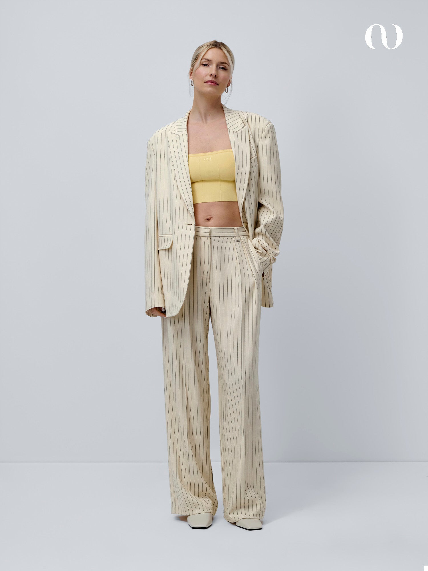 Eine Person steht vor einem schlichten hellen Hintergrund und trägt einen beigen Nadelstreifenanzug mit weit geschnittener Hose, einen übergroßen Blazer und das schmal geschnittene Juana Top von LeGer by Lena Gercke in Gelb. Ihr blondes Haar ist zurückgebunden, während sie selbstbewusst in die Kamera schaut.