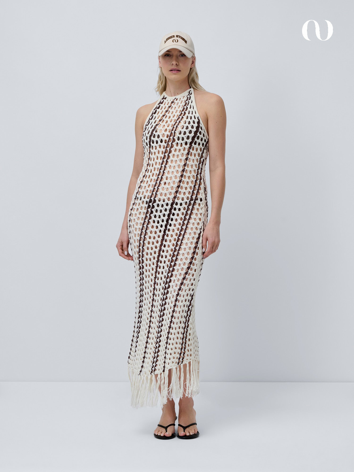 Eine Person steht vor einem schlichten Hintergrund und trägt das Salma Dress von LeGer by Lena Gercke - ein ärmelloses 7/8-langes Häkelkleid mit schwarzen und cremefarbenen Streifen, Fransen am Saum, beiger Kappe und schwarzen Sandalen für einen perfekten Strandlook.