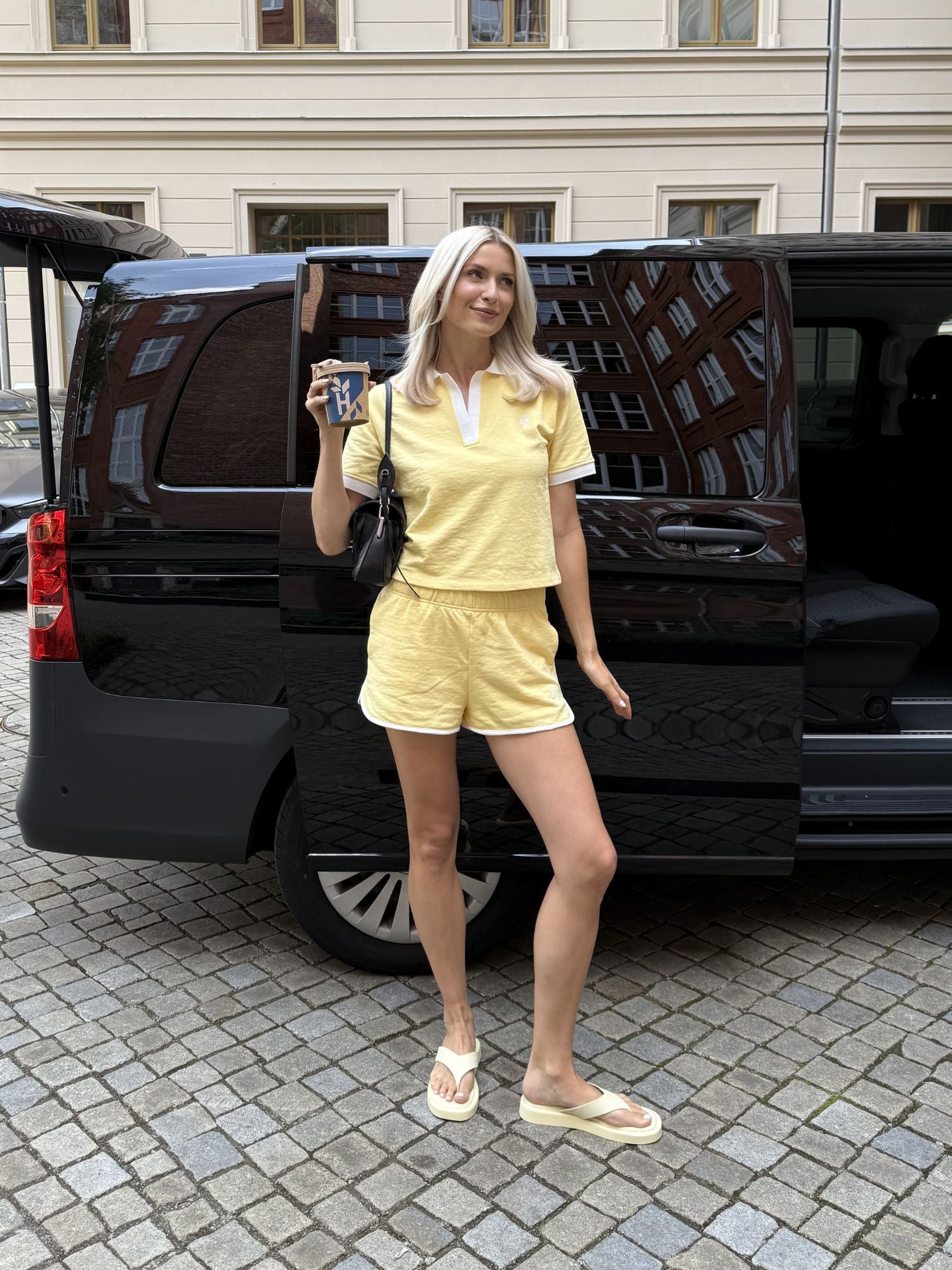 Eine blonde Frau in Größe 36 lächelt in die Kamera. Sie trägt ein gelbes Drew-T-Shirt von LeGer by Lena Gercke mit passenden Shorts und steht an einer offenen schwarzen Van-Tür, in der Hand ein Telefon und eine Kaffeetasse.