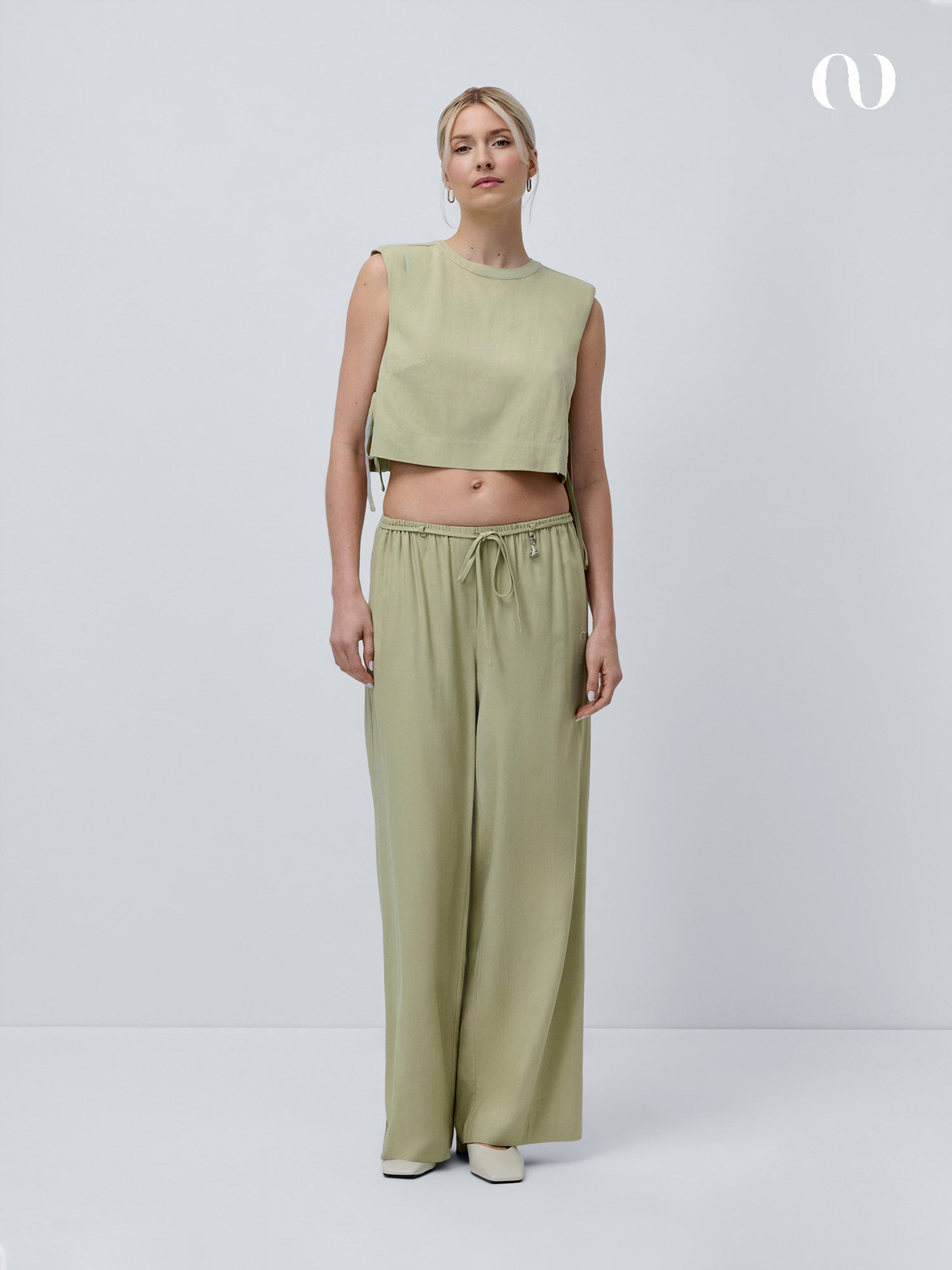 Eine Frau mit hellen, nach hinten gebundenen Haaren und Ohrringen trägt das mintgrüne Hanke Top von LeGer by Lena Gercke und eine dazu passende Hose mit weitem Bein und Kordelzug in der Taille vor einem schlichten hellen Hintergrund. Sie vervollständigt den Look mit weißen Schuhen.