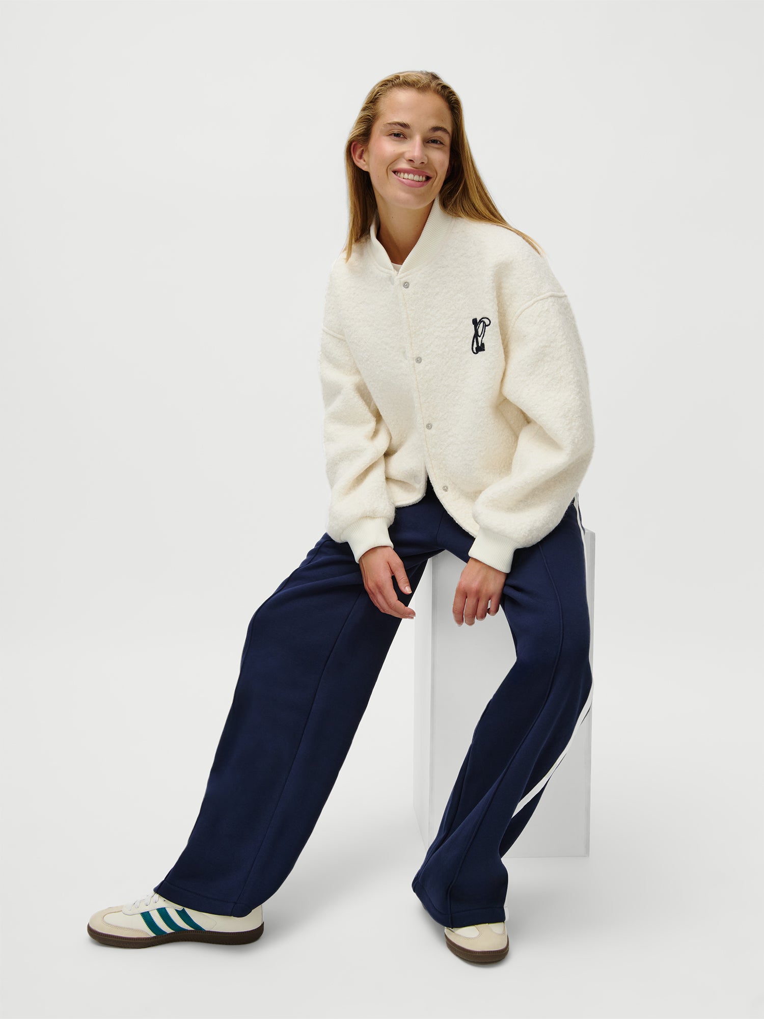 Eine lächelnde Person mit langen Haaren sitzt auf einem weißen Block und trägt die cremefarbene LeGer x Oh April - Blanca Jacket von LeGer by Lena Gercke, eine marineblaue Hose mit weitem Bein und weiße Turnschuhe mit grünen Streifen. Der Hintergrund ist einfarbig weiß.