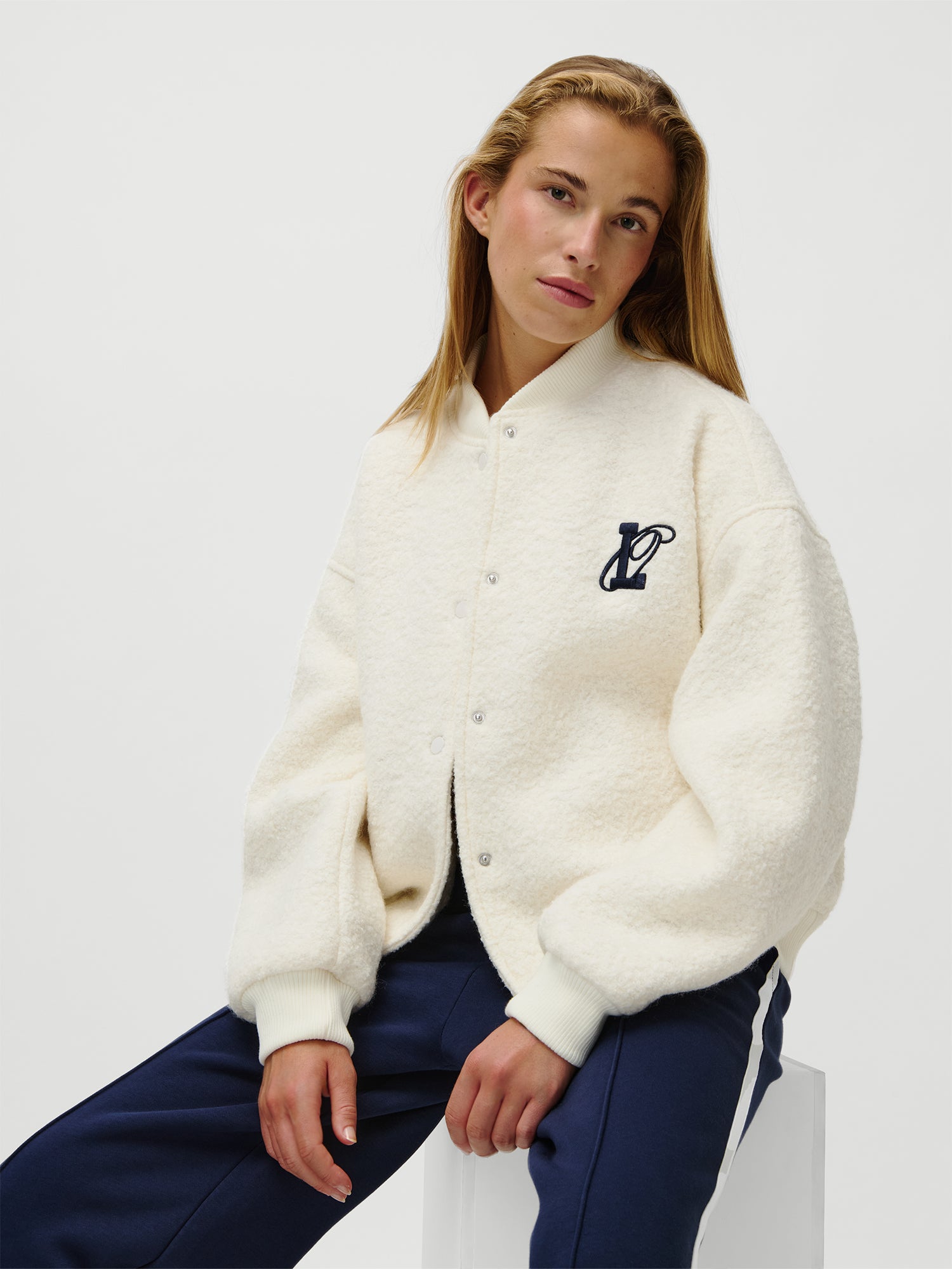 Eine Person mit langen blonden Haaren trägt die LeGer x Oh April - Blanca Jacket von LeGer by Lena Gercke, ein strukturiertes weißes Button-up in normaler Länge mit einem dunklen Brustlogo, gepaart mit einer marineblauen Hose, vor einem schlichten weißen Hintergrund.