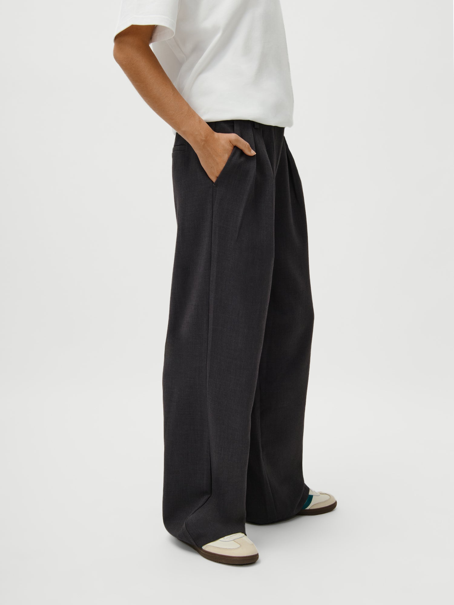 Model 1,73m trägt Größe 36: Franja Pants von LeGer by Lena Gercke in Schwarz (weit, lang/maxi), weiße Kurzarmbluse und weiße Sneaker mit beige-grünen Akzenten, stehend mit einer Hand in der Tasche vor weißem Hintergrund.