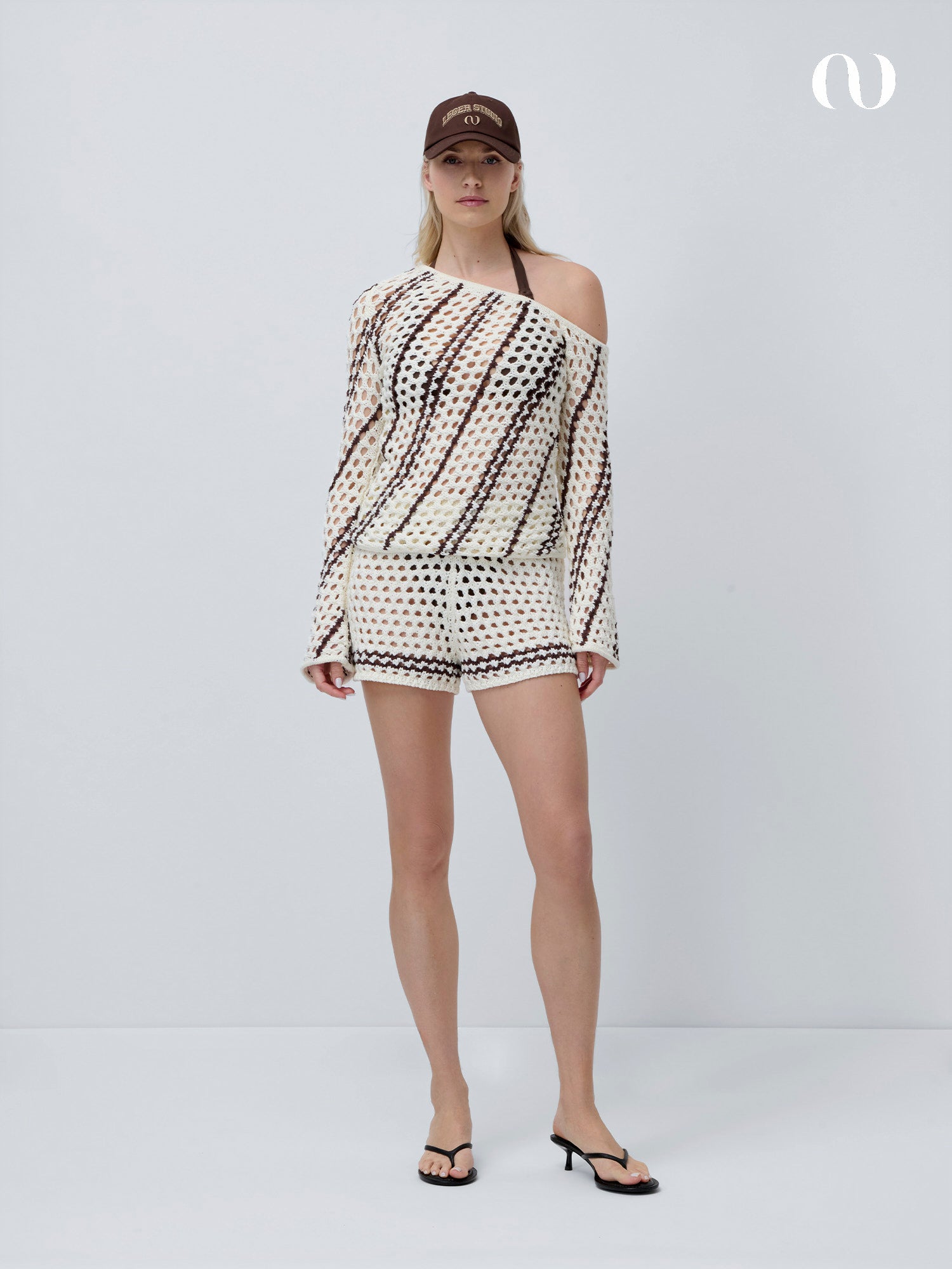 Eine Frau trägt die Laren Shorts von LeGer by Lena Gercke und ein dazu passendes Häkeltop, gestylt mit einem schwarzen Bikinioberteil, schwarzen Flip-Flops und einer braunen Baseballmütze, vor einem schlichten weißen Hintergrund.