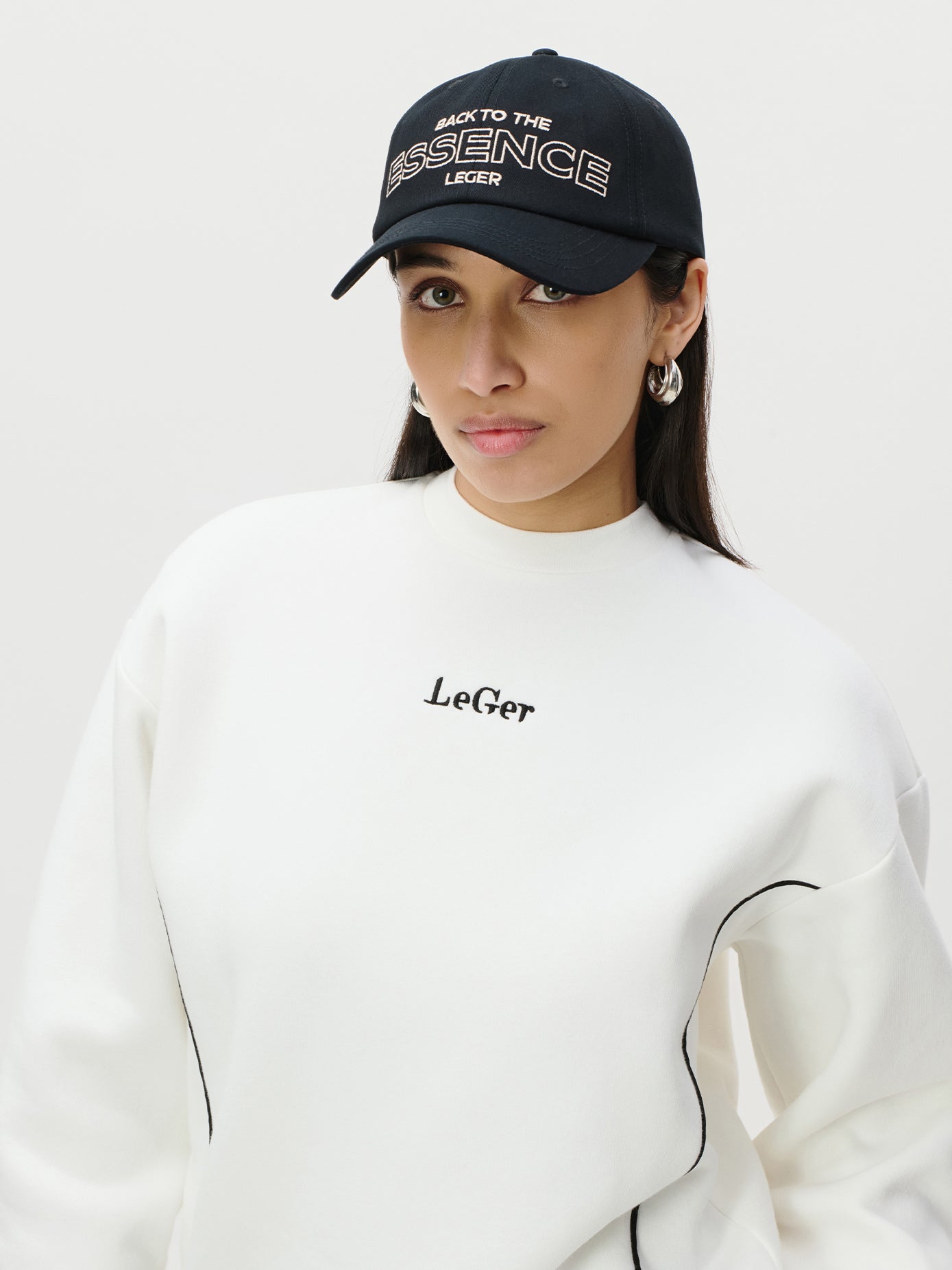 Eine Frau mit einer schwarzen "Iris Cap" von LeGer by Lena Gercke und silbernen Ohrringen posiert in einem weißen "LeGer"-Sweatshirt vor einem schlichten Hintergrund. Weitere Details finden Sie in der Produktinformation.