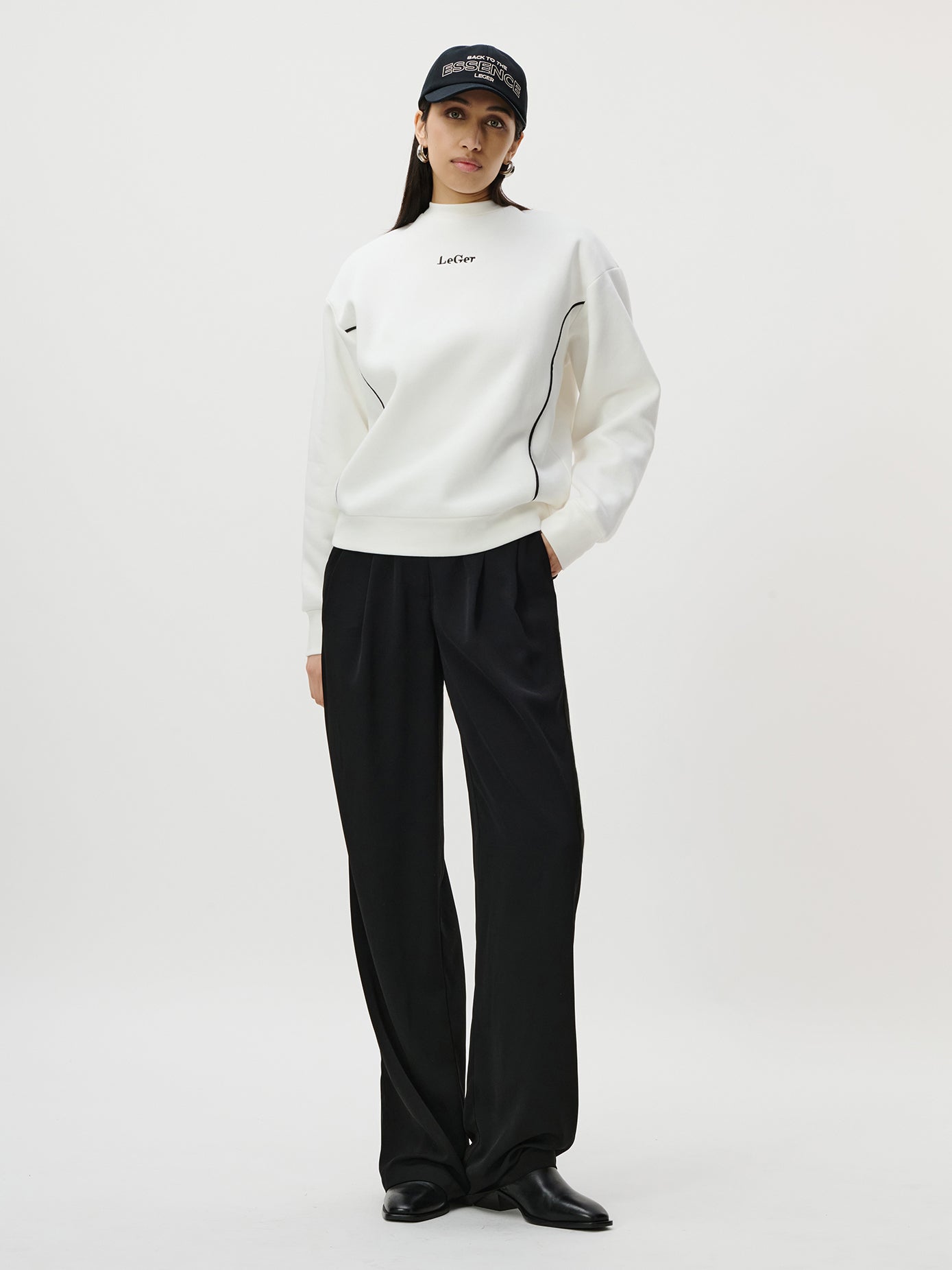 Vor einem schlichten Hintergrund steht eine Frau, die ein weißes Sweatshirt mit der Aufschrift „Letter“ und Produktinformationen, eine schwarze Hose mit weitem Bein, schwarze Schuhe und die Iris-Kappe von LeGer by Lena Gercke trägt. Ihre Hände stecken in den Taschen.