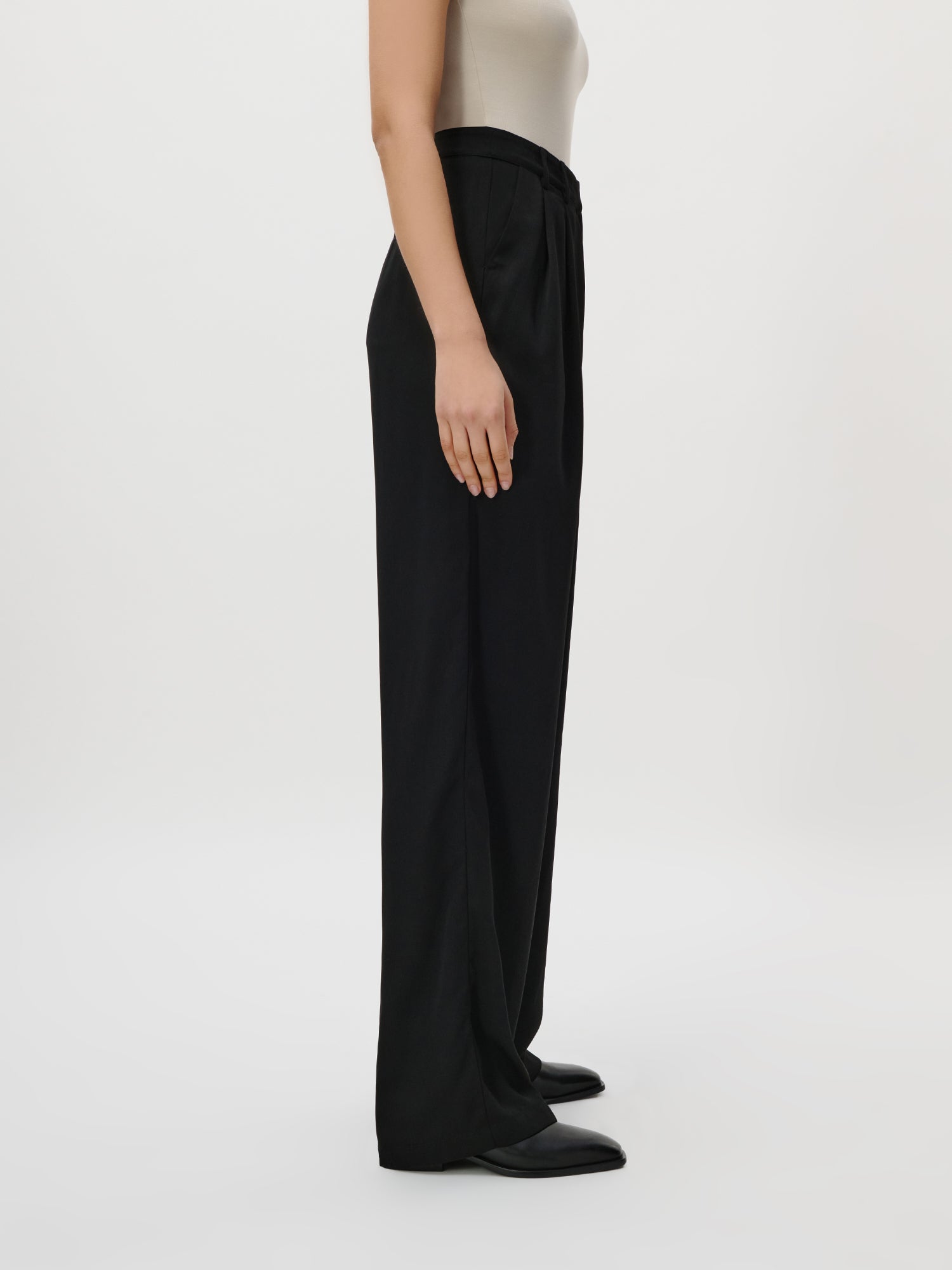 Eine Person (Größe 36, Model 1,77m) steht im Profil vor einem einfarbig weißen Hintergrund und trägt die Kora Pants Tall von LeGer by Lena Gercke - eine hochtaillierte, weite schwarze Hose - kombiniert mit einem ärmellosen beigen Top und schwarzen Schuhen. Nur die Schultern sind sichtbar.