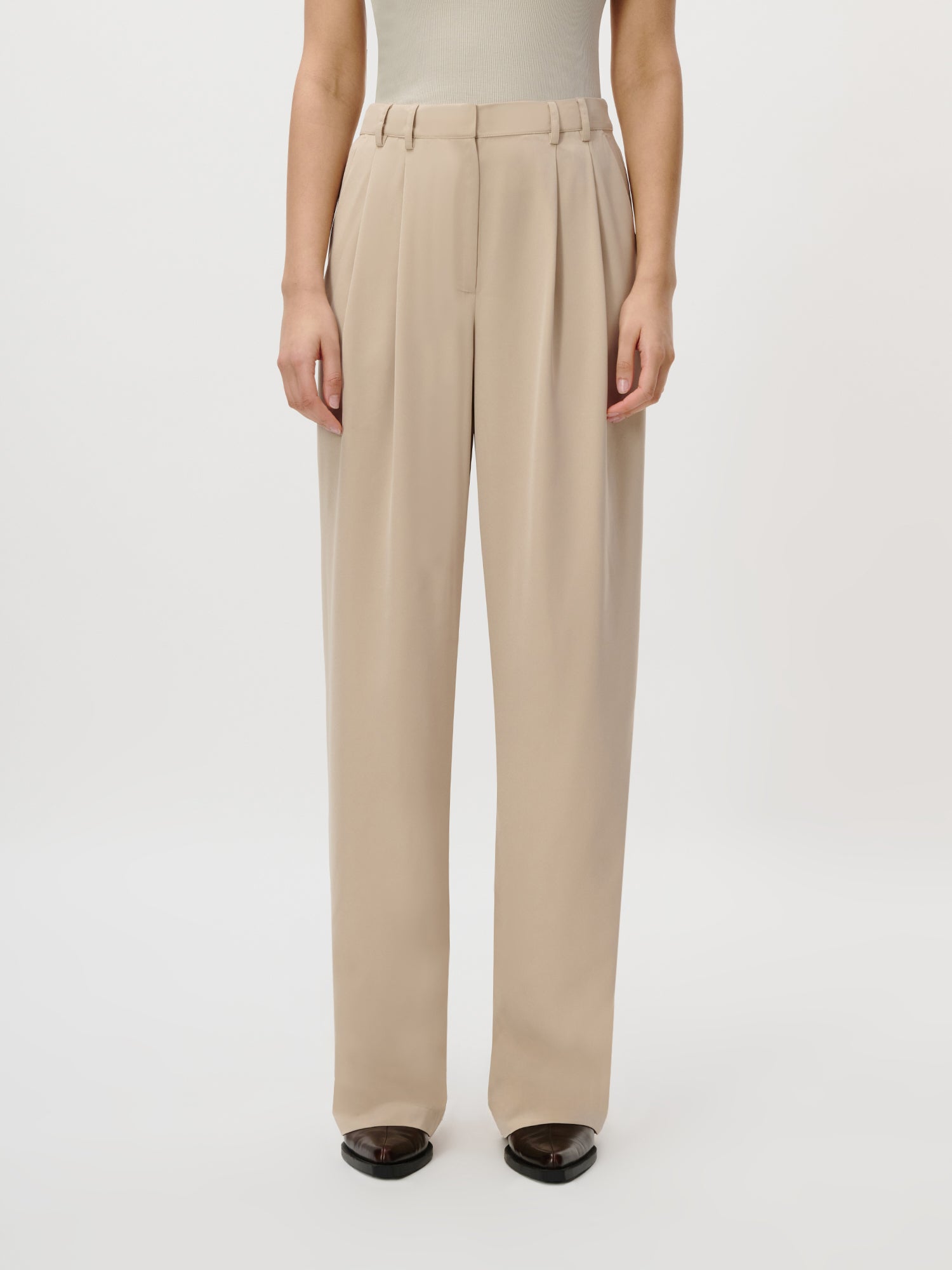 Eine Person trägt die Kora Pants Tall von LeGer by Lena Gercke in Beige (Größe 36) mit hoher Taille und weitem Bein mit Bundfalten, gestylt mit einem hellen Oberteil und schwarzen Schuhen, vor einem schlichten weißen Hintergrund.