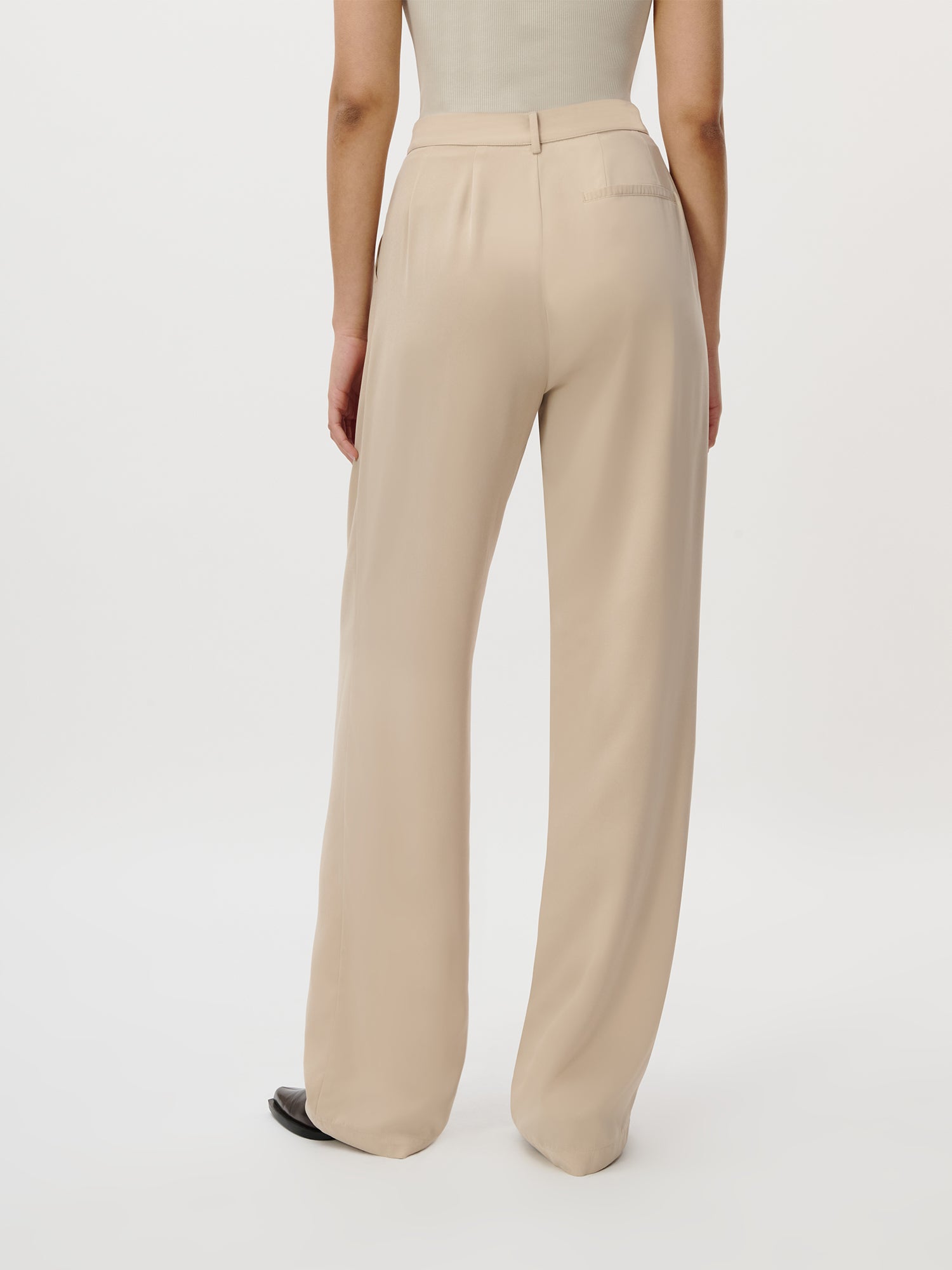 Eine Person (1,77m, Größe 36) trägt die LeGer by Lena Gercke Kora Pants Tall - eine hochtaillierte, locker sitzende beigefarbene Hose - mit einem leichten ärmellosen Oberteil und steht mit dem Rücken zur Kamera vor einem schlichten weißen Hintergrund.