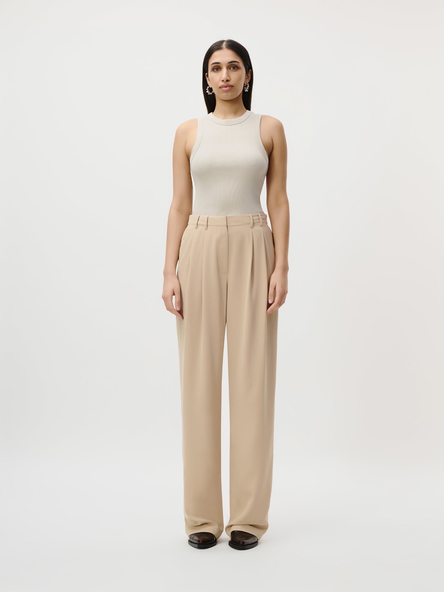 Eine Frau blickt vor einem schlichten weißen Hintergrund nach vorne und trägt die Kora Pants Tall von LeGer by Lena Gercke in Größe 36 - eine beigefarbene Hose mit hoher Taille und weitem Bein - kombiniert mit einem ärmellosen beigen Top, schwarzen Schuhen und goldenen Ohrringen.
