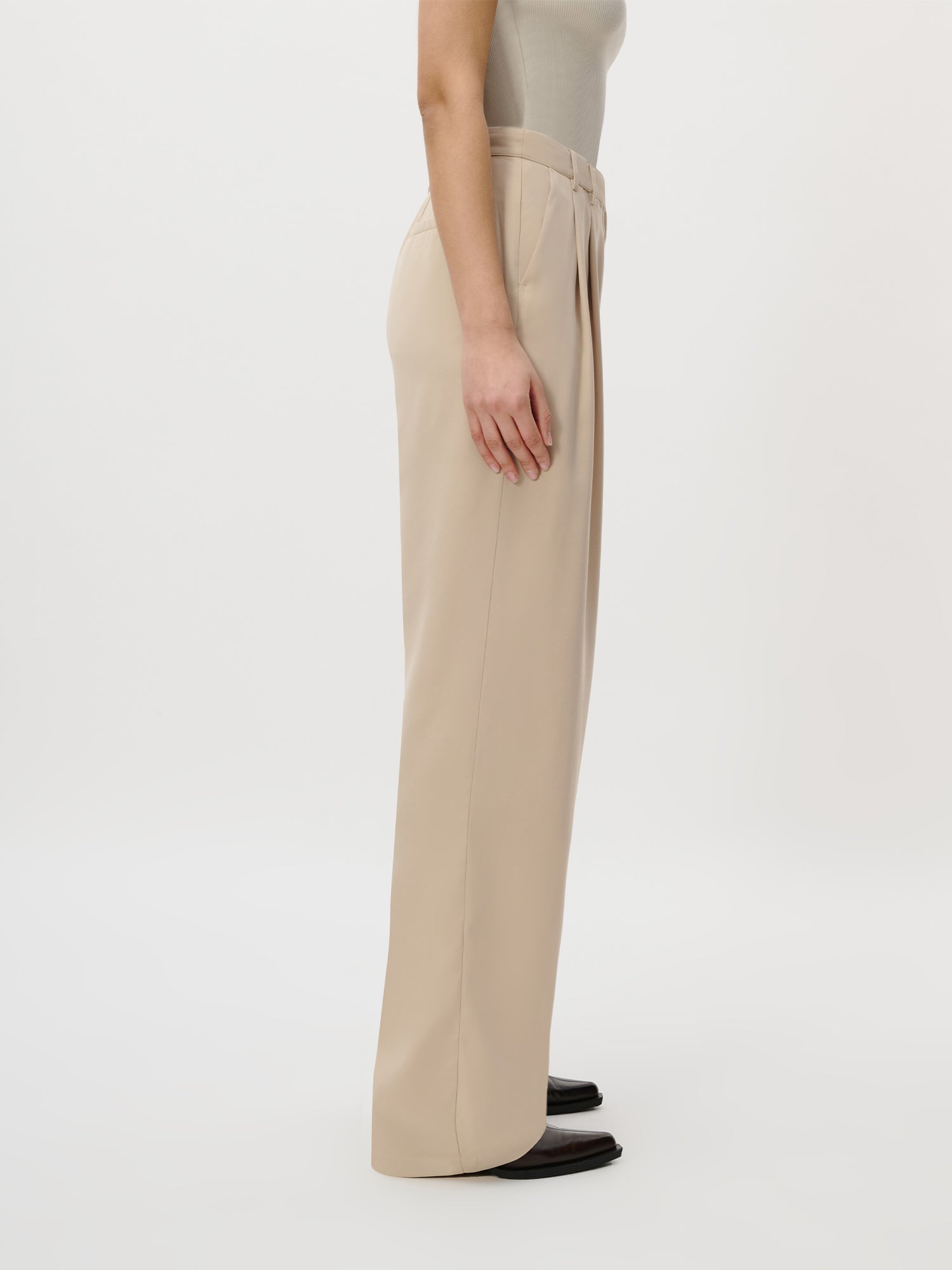 Eine Person im Profil modelliert die Kora Pants Tall von LeGer by Lena Gercke in Beige, gepaart mit einem leichten ärmellosen Top und schwarzen Schuhen in Größe 36, vor einem schlichten weißen Hintergrund.