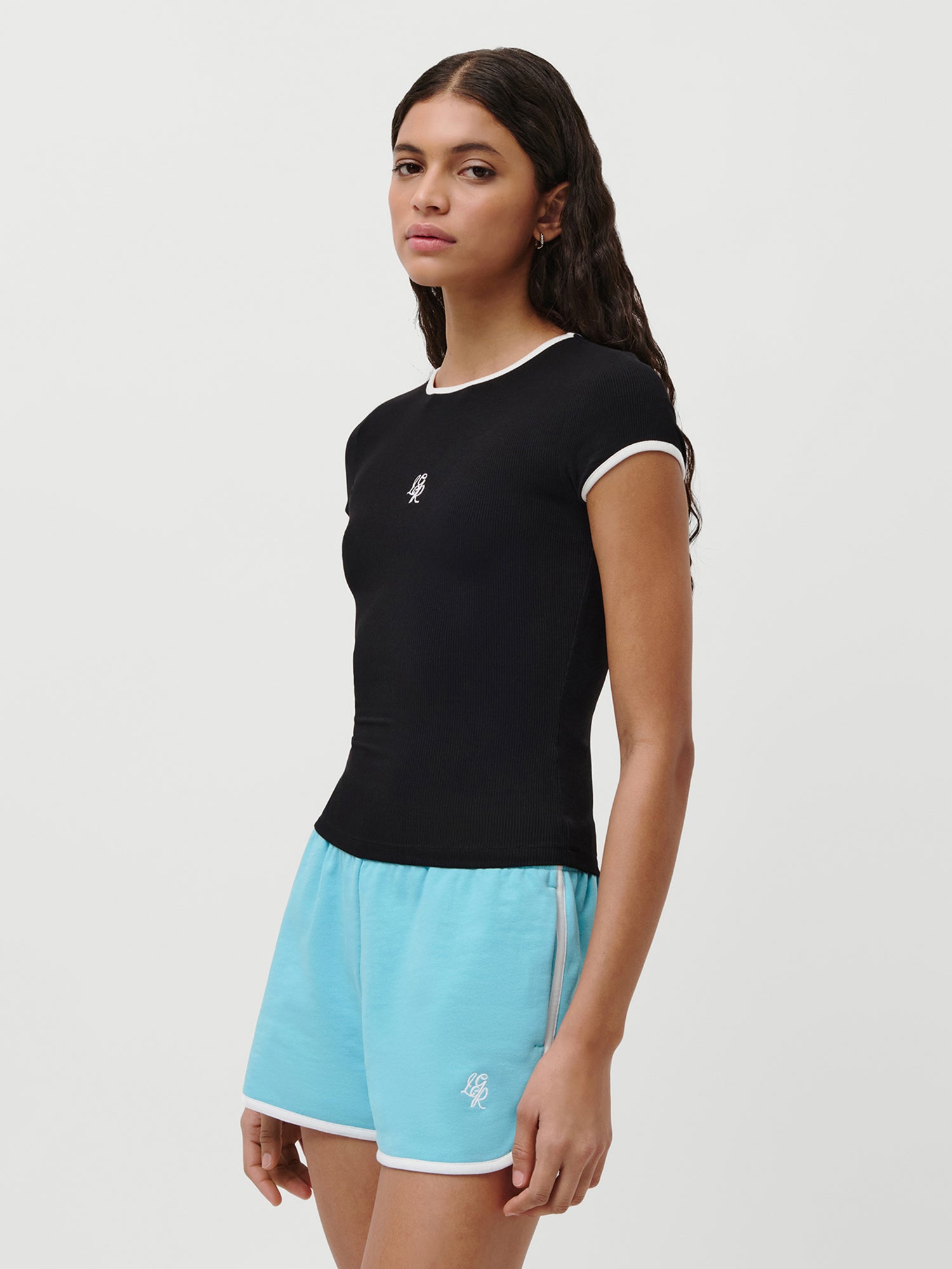 Eine Frau mit langen, gewellten dunklen Haaren trägt das LeGer by Lena Gercke Elmira T-Shirt in Schwarz mit weißem Rand und hellblaue Sporthosen. Sie steht vor einem schlichten weißen Hintergrund und blickt leicht zur Seite.