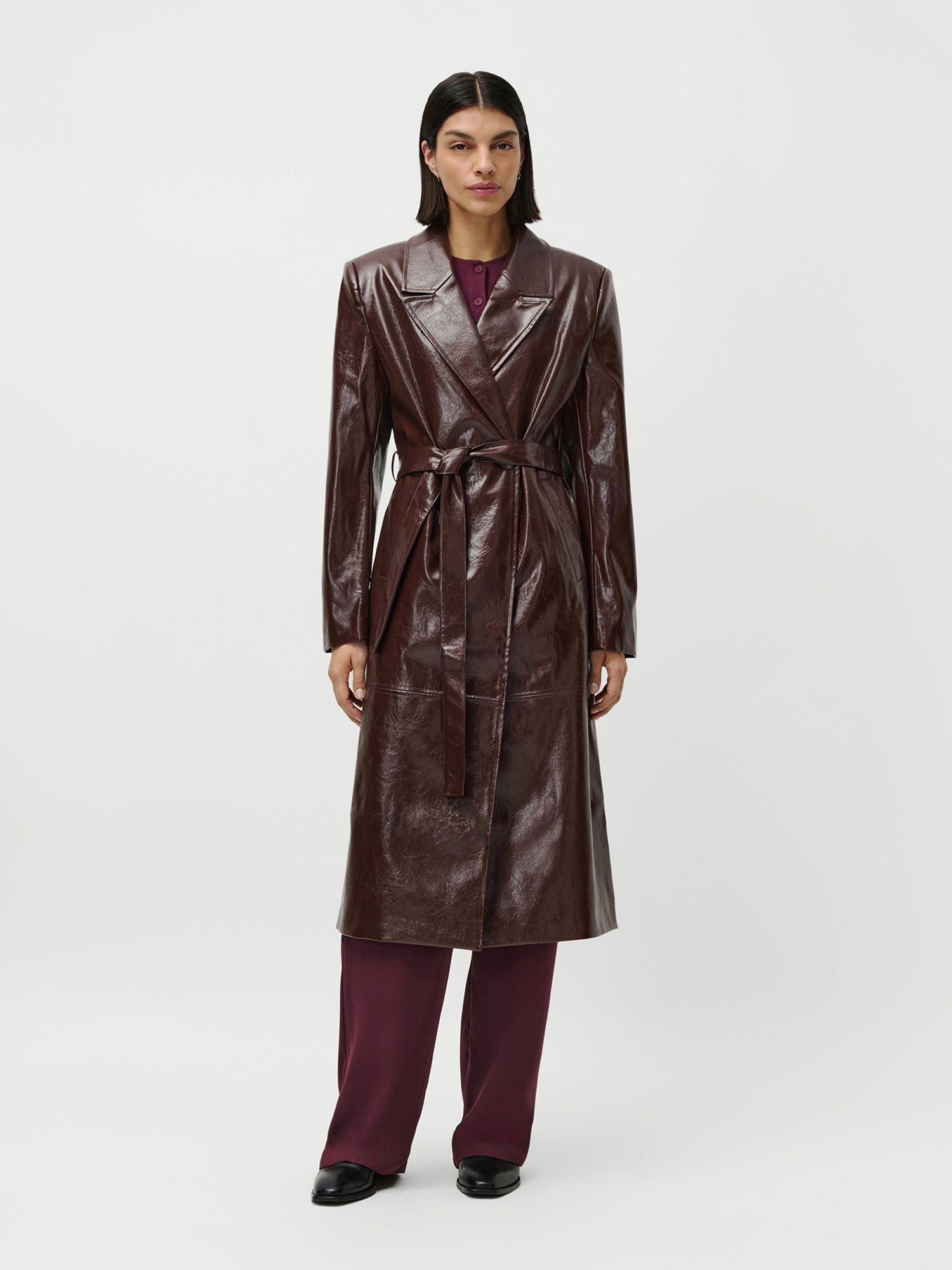 Eine Person steht mit dem Gesicht nach vorne und trägt den LeGer by Lena Gercke Frieda Coat - einen langen, dunkelbraunen Ledermantel mit Gürtel - über einer burgunderroten Hose und einem passenden Oberteil mit normaler Passform, dazu schwarze Schuhe vor einem schlichten weißen Hintergrund.