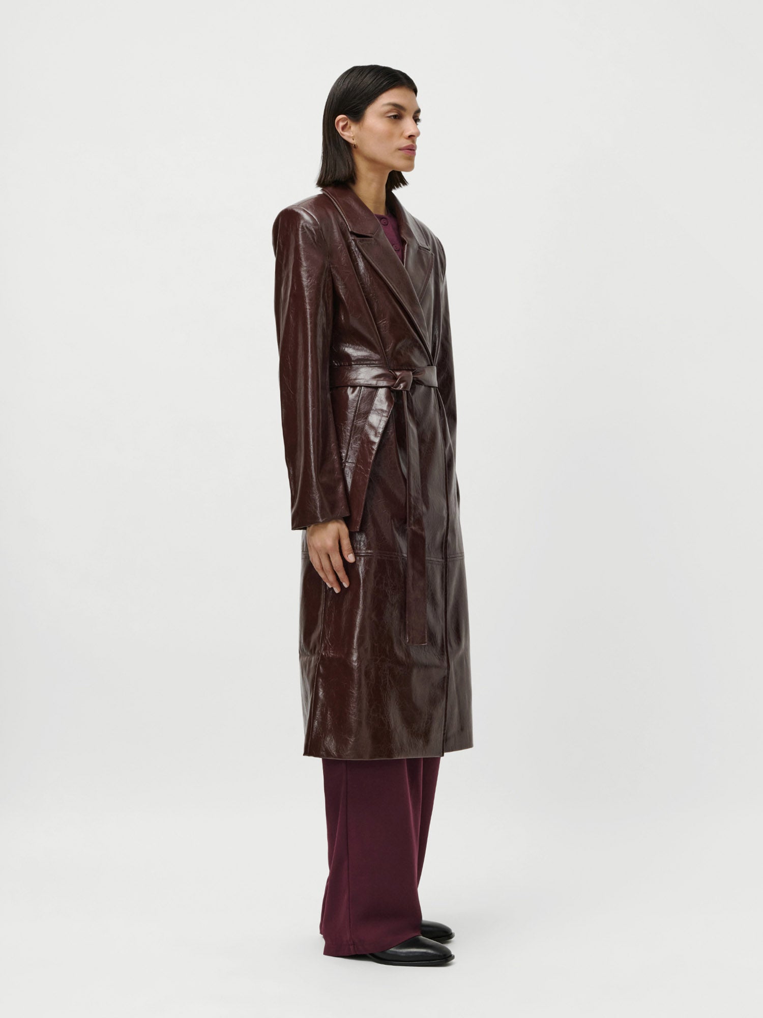 Vor einem schlichten weißen Hintergrund steht eine Person im LeGer by Lena Gercke Frieda Coat - einem langen, dunkelbraunen Ledermantel mit Gürtel und langen Ärmeln - über einer weinroten Hose und schwarzen Schuhen, die dunklen glatten Haare hinter die Ohren gesteckt.