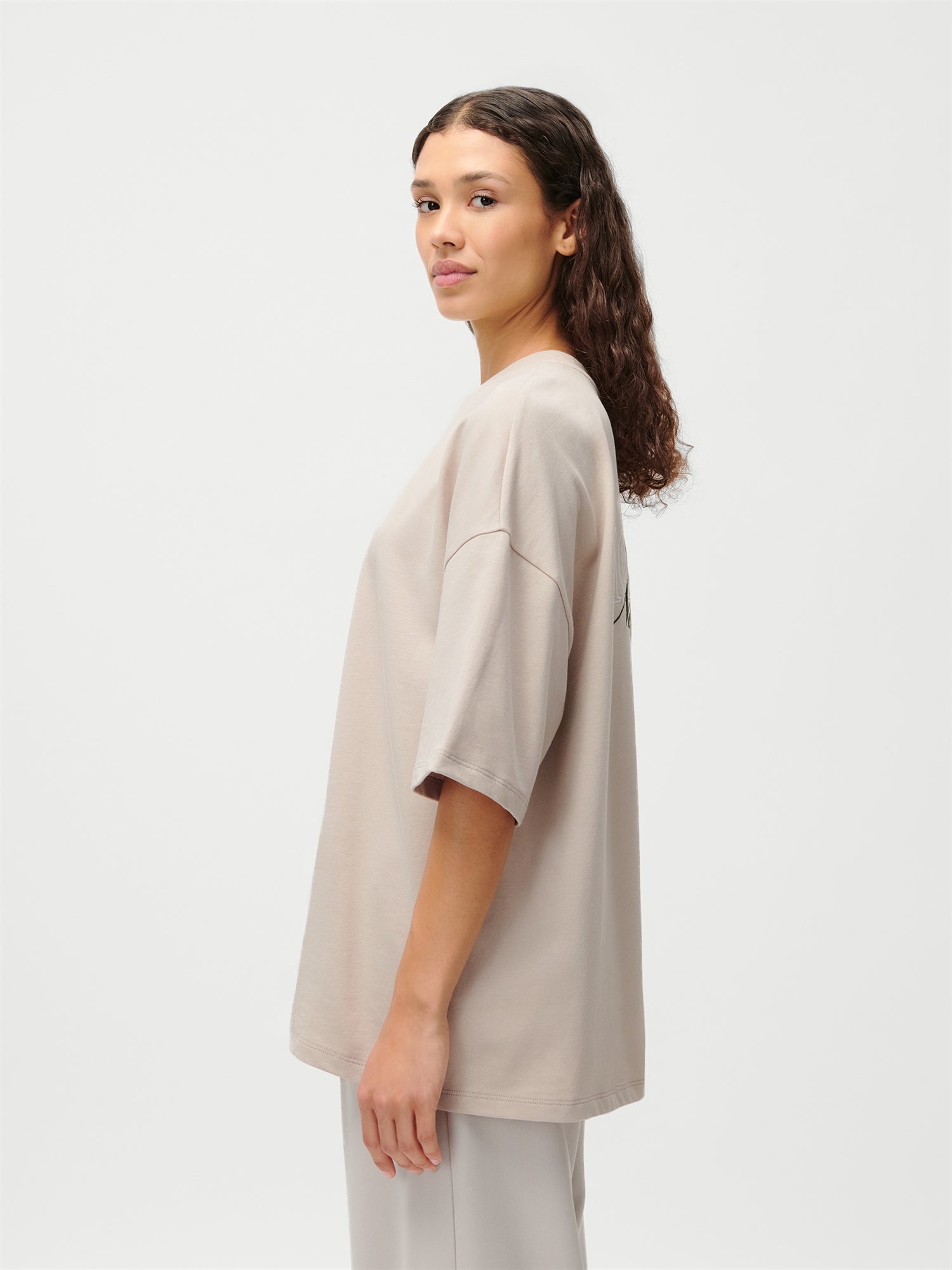 Eine Frau mit gewelltem braunem Haar steht seitlich und schaut in die Kamera. Sie trägt das beigefarbene Suzi T-Shirt in Übergröße von LeGer by Lena Gercke mit einer hellen Hose vor einem schlichten weißen Hintergrund.