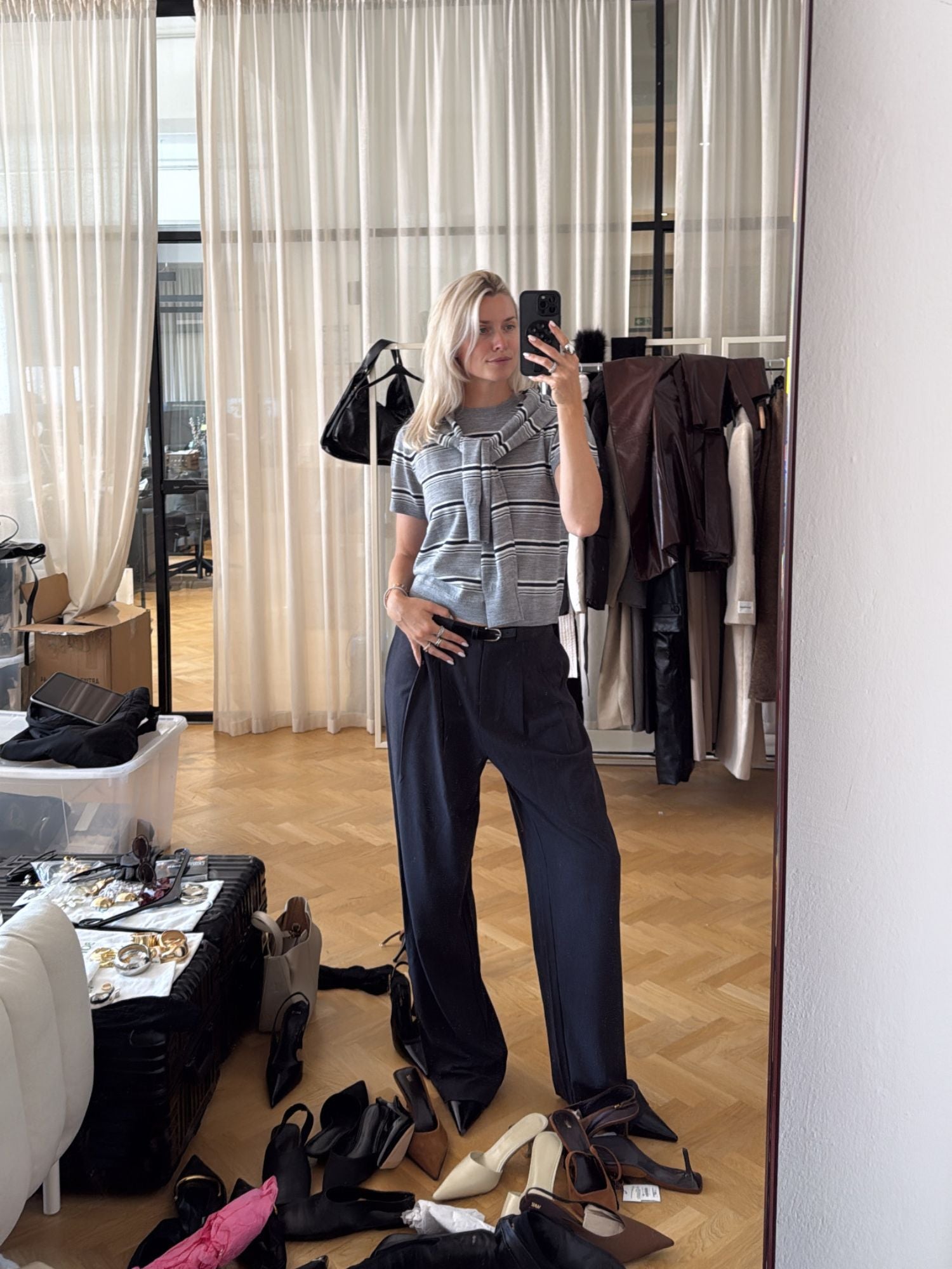 Eine Frau macht ein Spiegel-Selfie in einem stilvollen Büro. Sie trägt den Selma-Pullover von LeGer by Lena Gercke und eine Hose mit weitem Bein. Kleidung, Schuhe und Accessoires sind auf dem Boden und den Möbeln um sie herum verstreut.