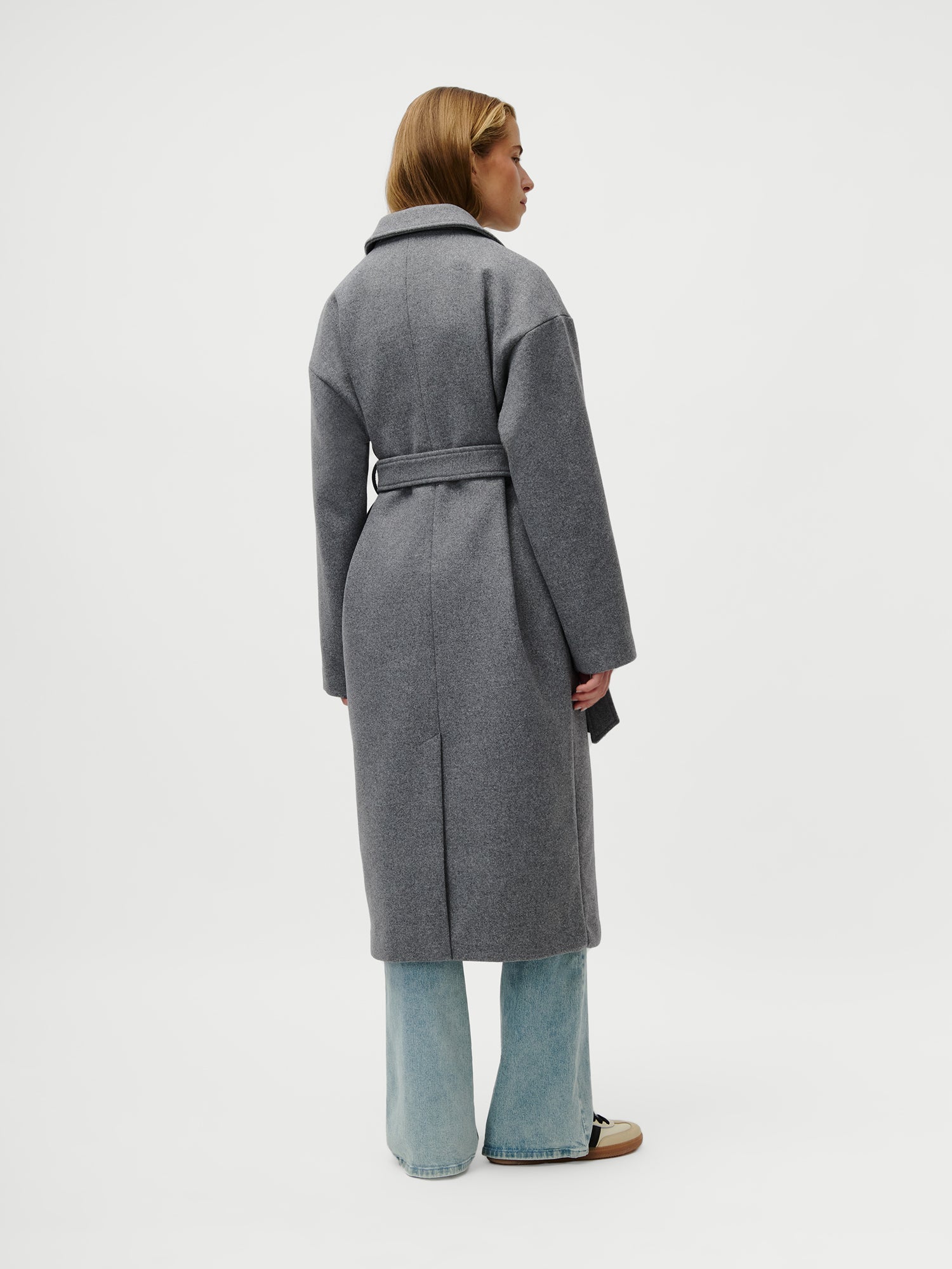 Eine Person blickt weg und trägt den Anaida Coat von LeGer by Lena Gercke - einen langen, grauen, locker sitzenden Mantel mit langen Ärmeln - über hellblauen Jeans mit weitem Bein und beigen Turnschuhen vor einem schlichten weißen Hintergrund.