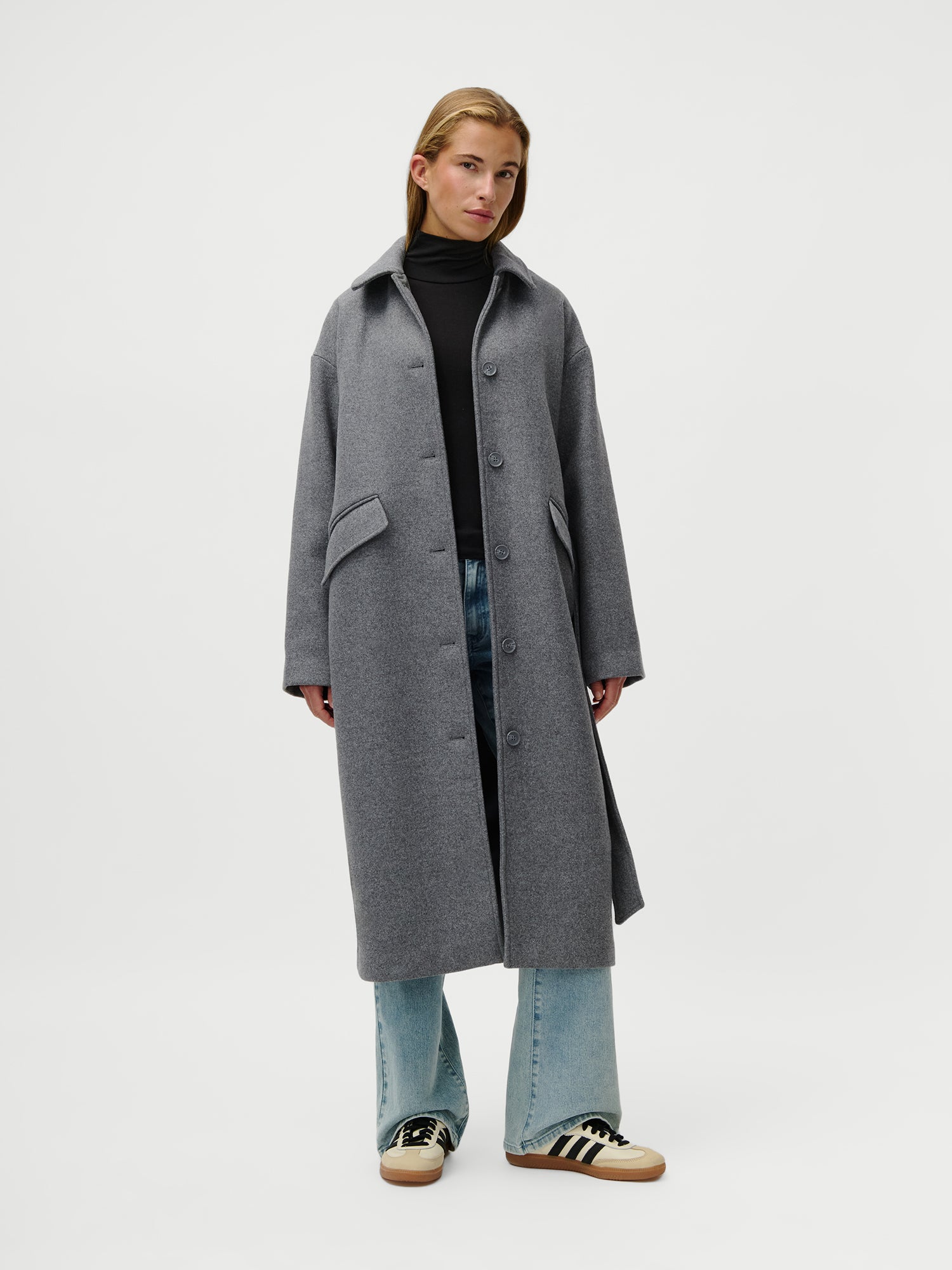 Eine Person steht vor einem weißen Hintergrund und trägt den Anaida Coat von LeGer by Lena Gercke, einen langen grauen Mantel in lockerer Passform, kombiniert mit einem schwarzen langärmeligen Rollkragenpullover, hellblauen Jeans und beigen Sneakers mit schwarzen Streifen.