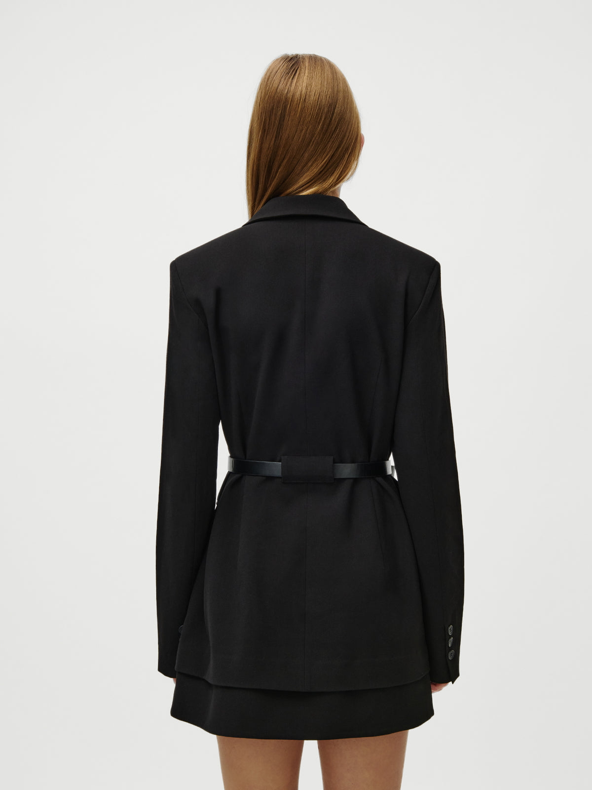 Eine Frau mit glattem, hellbraunem Haar ist von hinten zu sehen. Sie trägt das Kleid „Jeromina“ von LeGer by Lena Gercke, ein schwarz, normal geschnittenes Blazerkleid mit einem Gürtel in der Taille und Knopfdetails an den Ärmeln. Der Hintergrund ist einfarbig weiß.