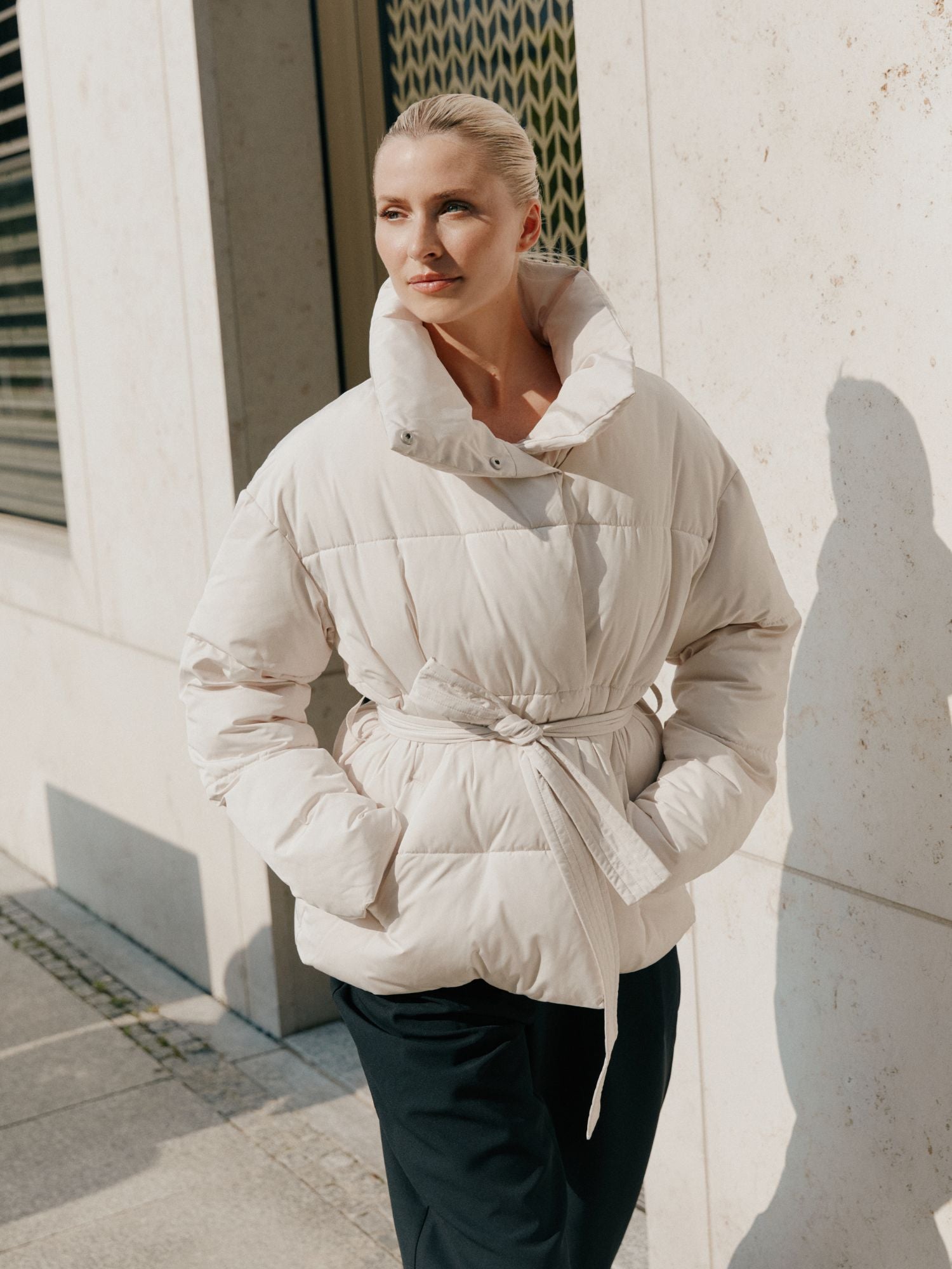 Eine blonde Frau mit zurückgebundenen Haaren trägt die cremefarbene Margitta Jacke von LeGer by Lena Gercke in Größe 36 mit langen Ärmeln und einem Gürtel, gepaart mit einer dunklen Hose, draußen vor einem hellen Steinhaus im Sonnenlicht.