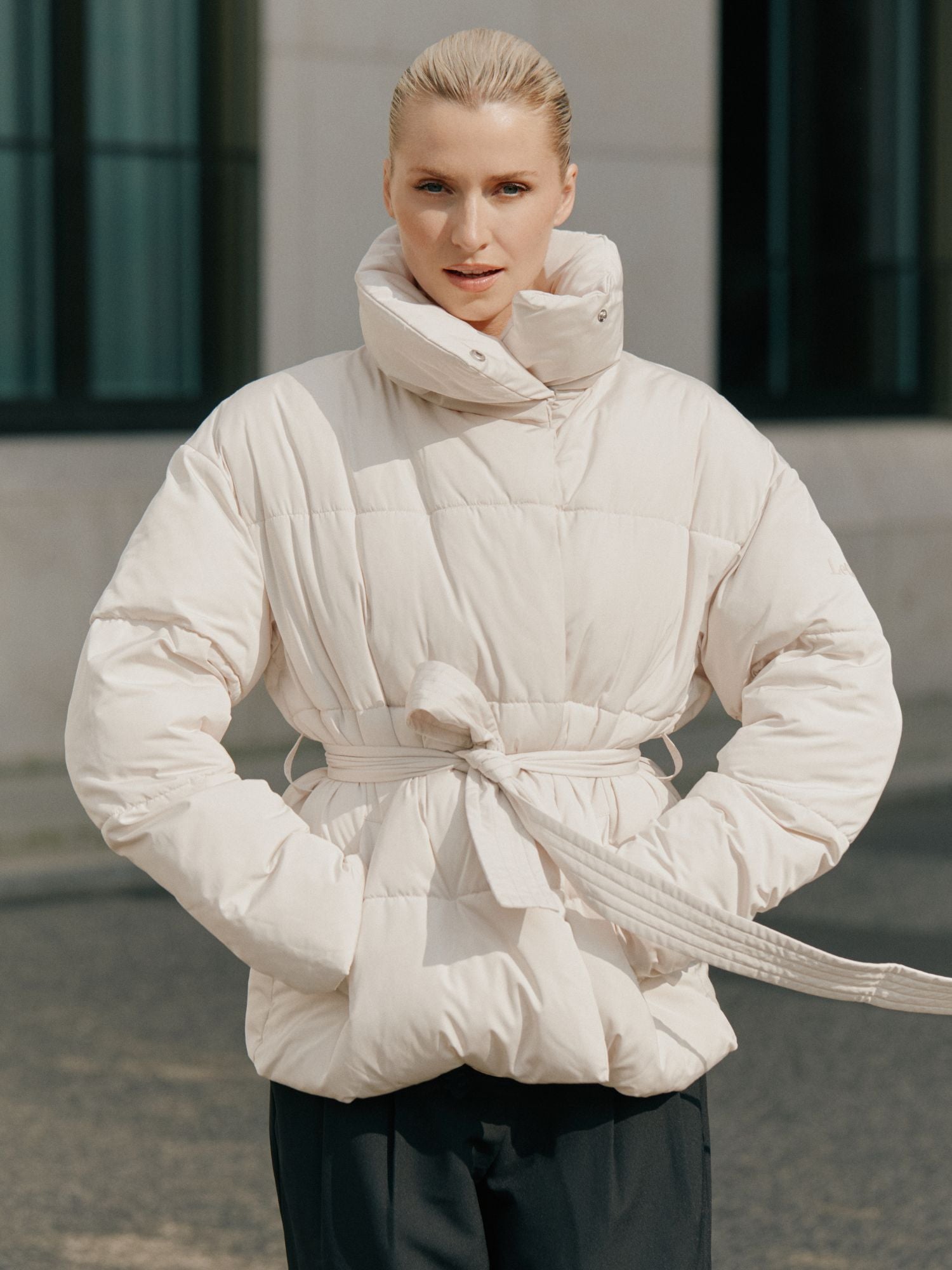 Eine Frau mit blondem Haar steht im Freien vor einem neutralen Gebäude und trägt die LeGer by Lena Gercke Margitta Jacket in Größe 36 - einen hellen, langärmeligen Puffer mit Gürtel und Taschen - und schaut in die Kamera.