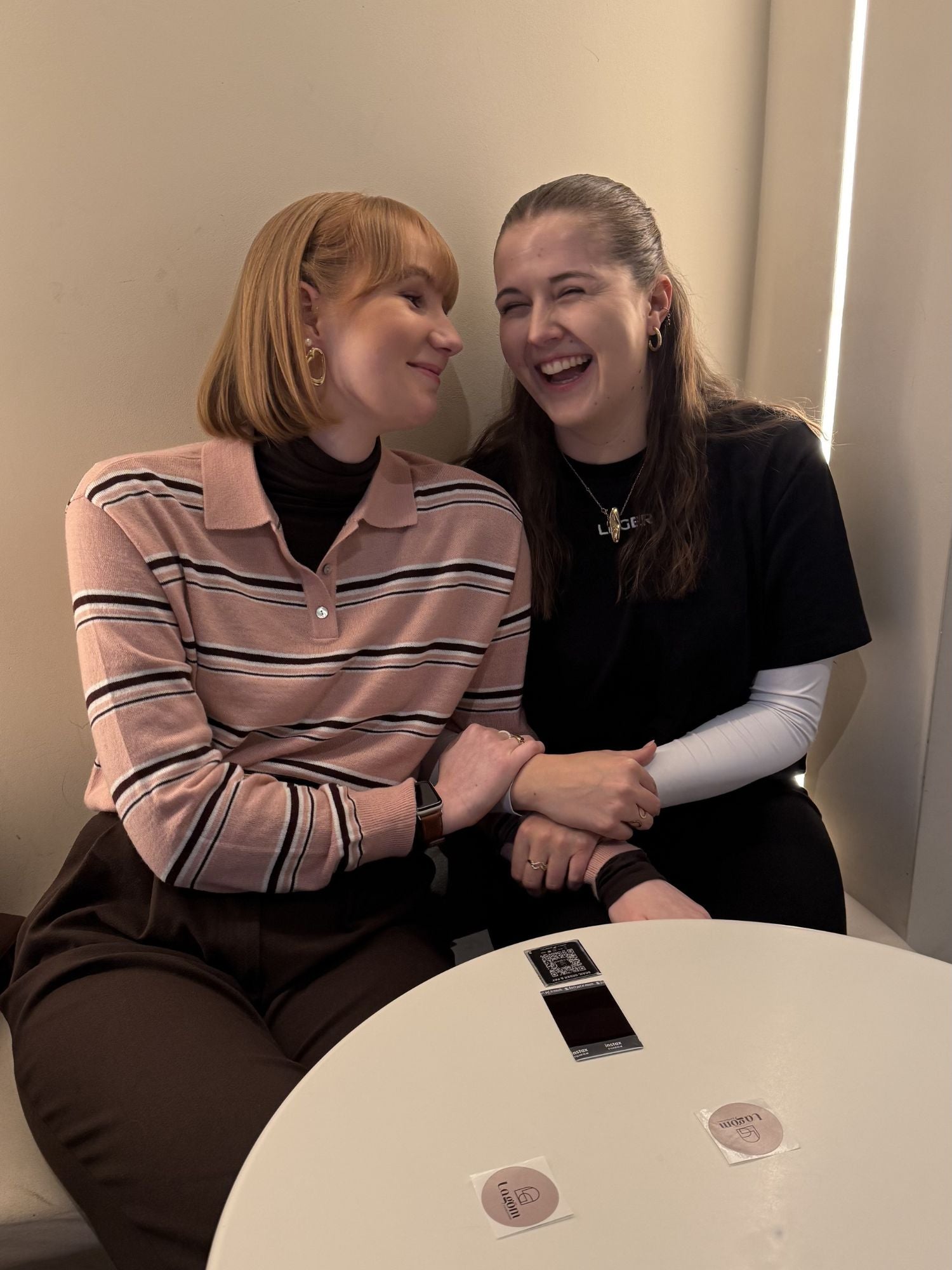 Zwei Frauen sitzen eng an einem runden weißen Tisch, lächeln und halten sich an den Händen. Die eine trägt den Lilith-Pullover von LeGer by Lena Gercke in Rosa, Größe 36, die andere ein schwarzes Shirt. Beide sehen glücklich und entspannt aus, vor ihnen stehen Getränke und Untersetzer.