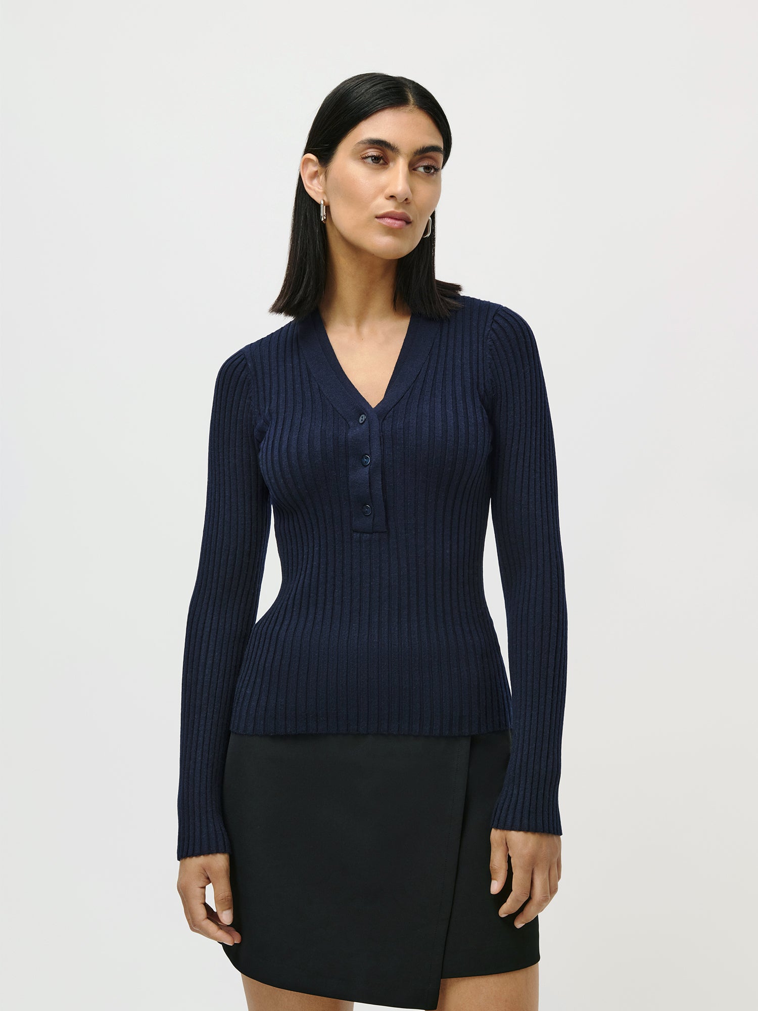 Eine Frau mit glattem dunklem Haar trägt das Nannette Longsleeve von LeGer by Lena Gercke - ein schmal geschnittenes, geripptes marineblaues Oberteil mit V-Ausschnitt, Knöpfen und normaler Länge - in Kombination mit einem schwarzen Rock vor einem schlichten hellen Hintergrund.