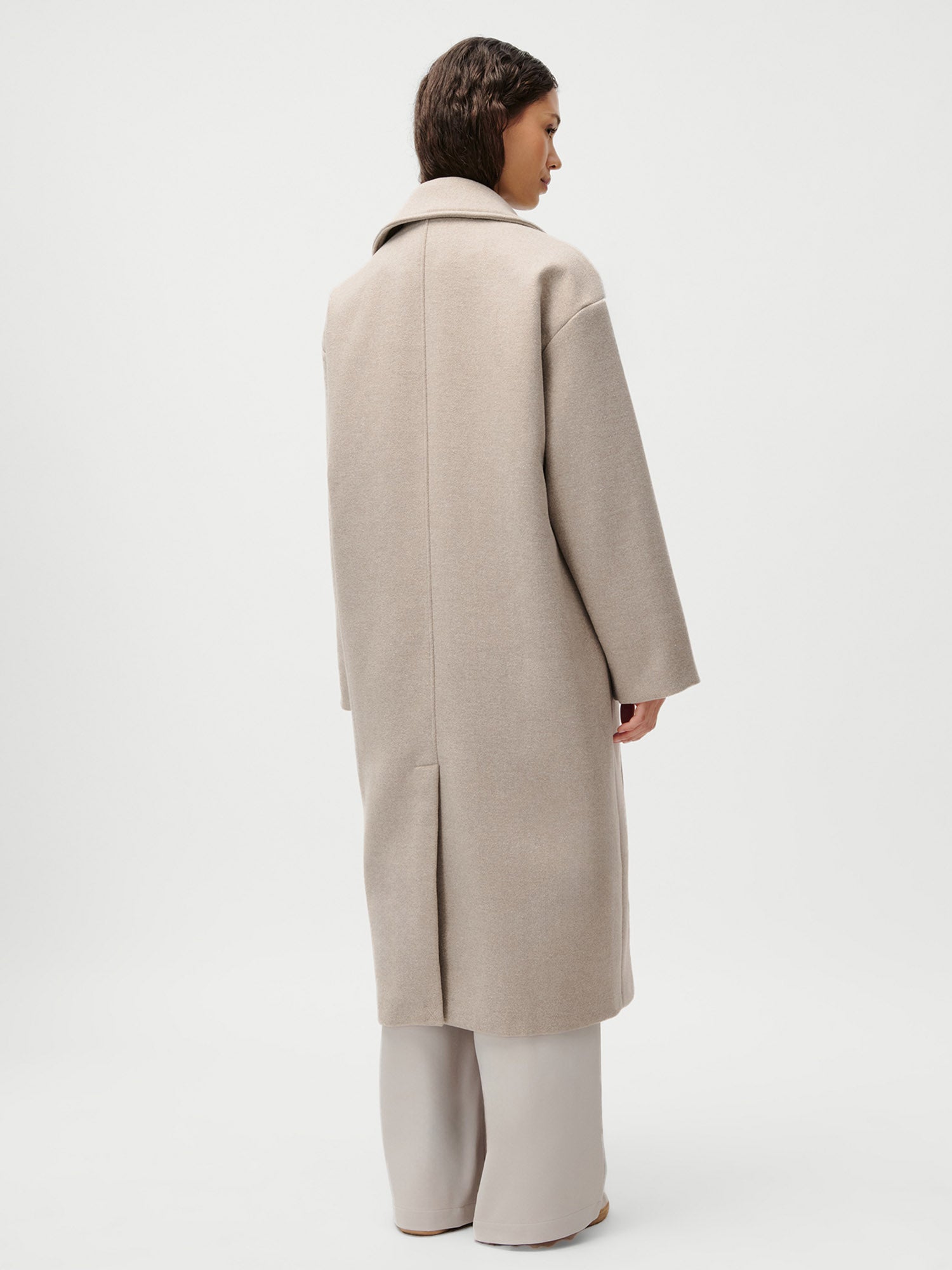Eine Person steht mit dem Gesicht zur Wand und trägt den Mantel Nelly von LeGer by Lena Gercke in Größe 36 mit Kragen und weiten Ärmeln, gepaart mit einer hellen Hose mit weitem Bein vor einem schlichten weißen Hintergrund.