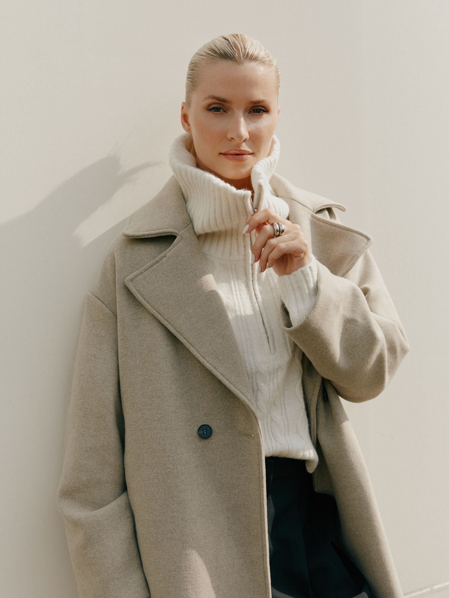 Eine Frau mit blondem Haar trägt den Nelly Coat von LeGer by Lena Gercke in Größe 36 über einem weißen Rollkragenpullover, steht vor einem hellen Hintergrund und berührt leicht ihren Kragen, während sie in die Kamera schaut.