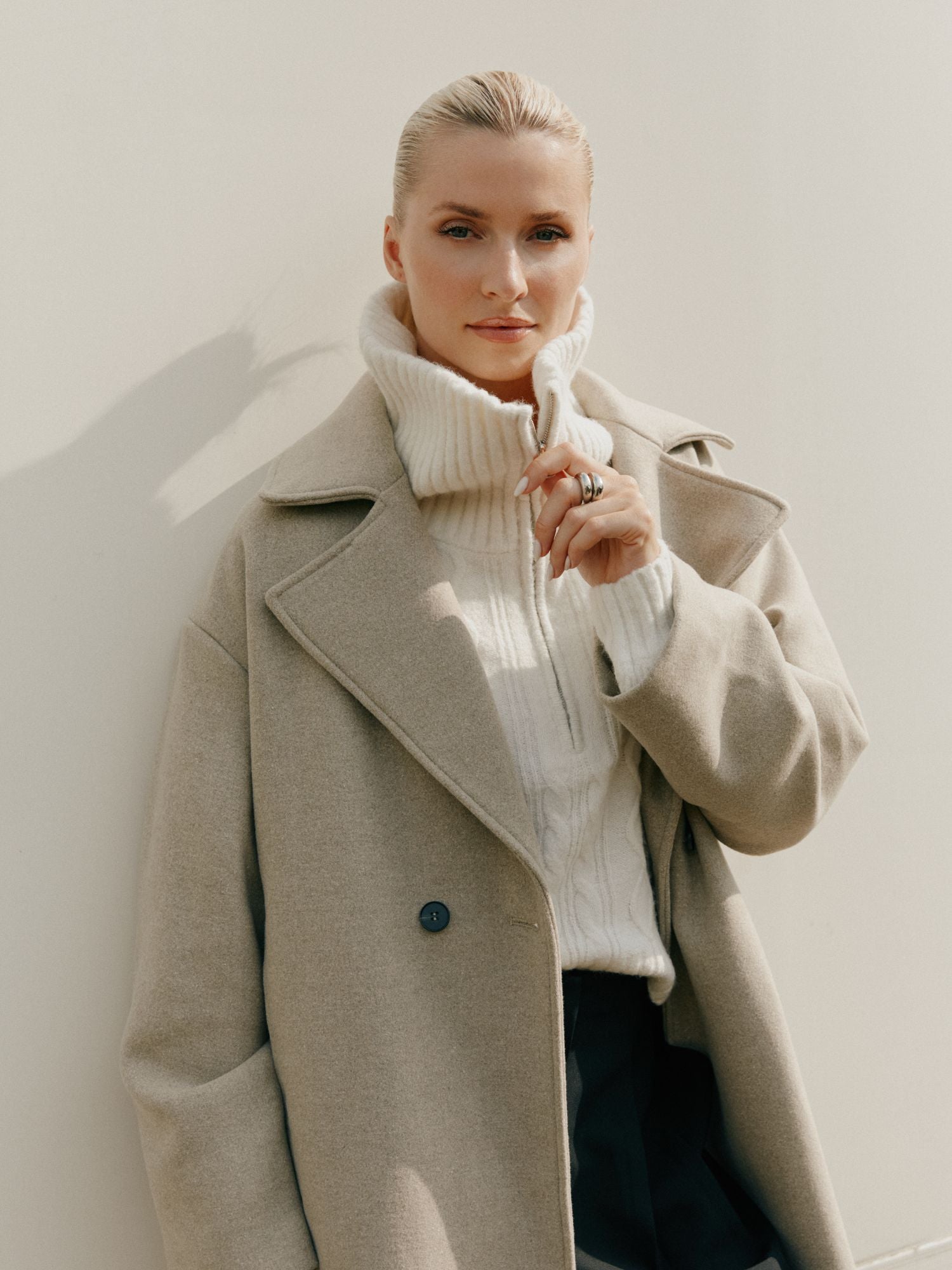 Eine blonde Frau mit hochgestecktem Haar trägt einen hellbeigen Mantel über dem Josefina-Pullover von LeGer by Lena Gercke. Sie steht vor einem schlichten hellen Hintergrund, berührt sanft ihren Kragen und schaut in die Kamera.