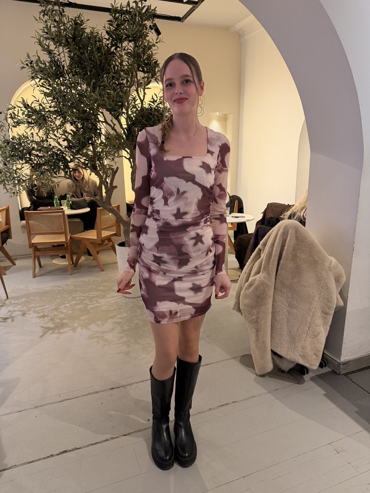 Eine Frau steht drinnen und lächelt in dem Loana-Kleid von LeGer by Lena Gercke - einem schmal geschnittenen, lila-weiß geblümten Kleid mit schwarzen kniehohen Stiefeln. Hinter ihr sitzen Menschen an Tischen neben einem großen, getopften Baum.