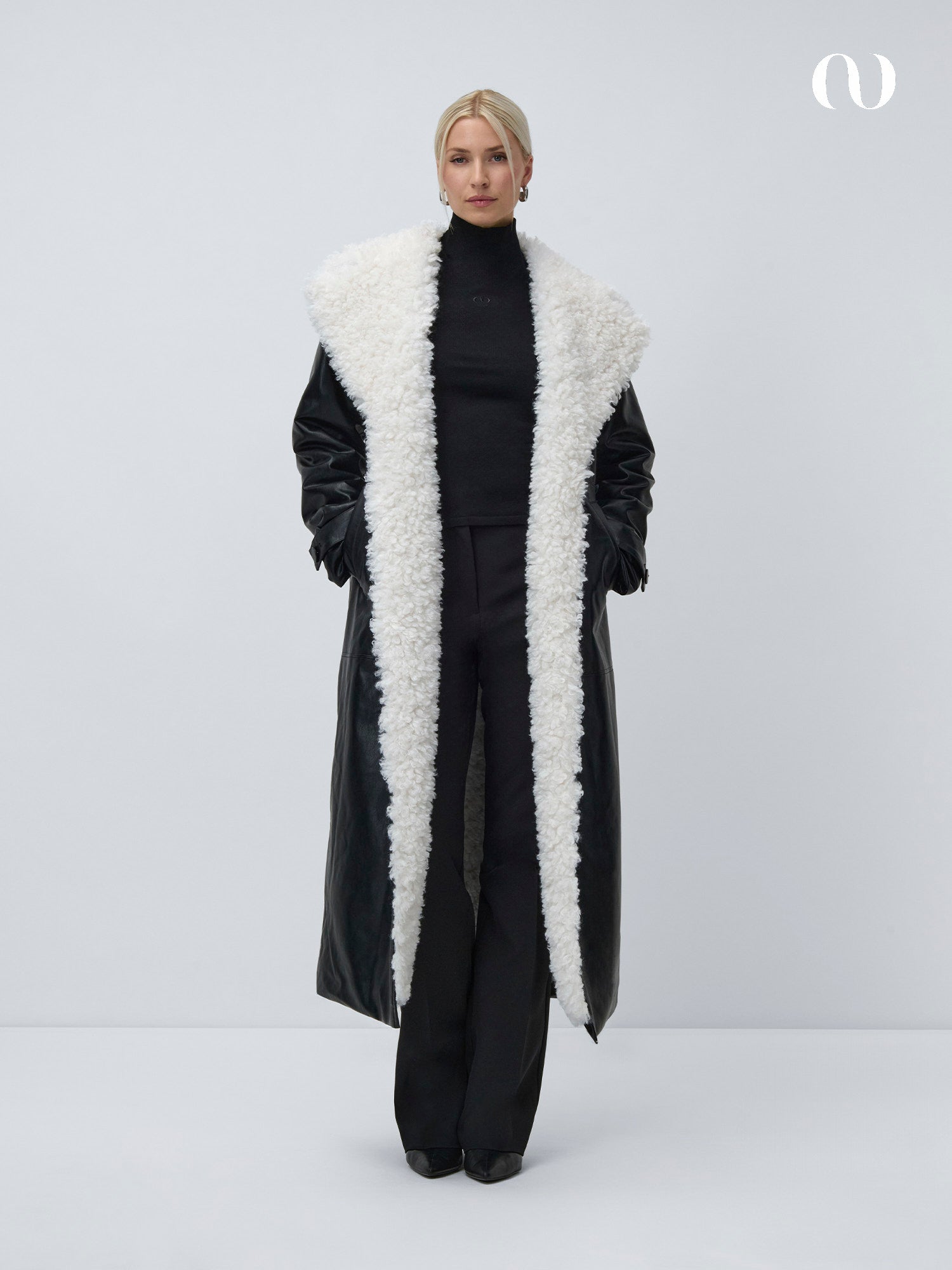 Eine Frau mit zurückgestyltem Haar und neutralem Gesichtsausdruck steht vor einem schlichten Hintergrund und trägt den eleganten Janique Coat von LeGer by Lena Gercke - einen langen Mantel mit einem großen, flauschigen weißen Kragen - über einem schwarzen Rollkragen und einer Hose.
