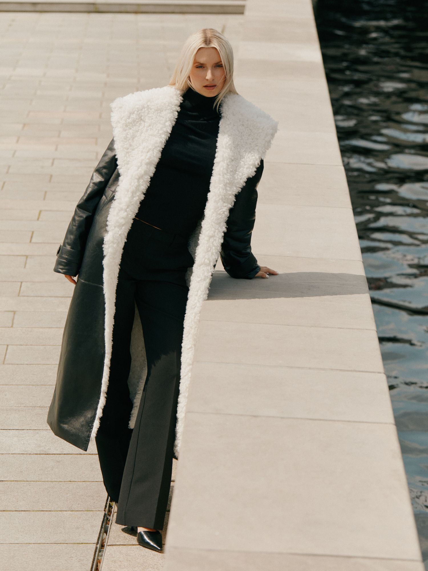 Eine blonde Frau lehnt an einem hellen Steinsims am Wasser und trägt den Janique Mantel von LeGer by Lena Gercke mit weißem Shearling-Besatz, gepaart mit einer schwarzen Hose und schwarzen spitzen Schuhen.