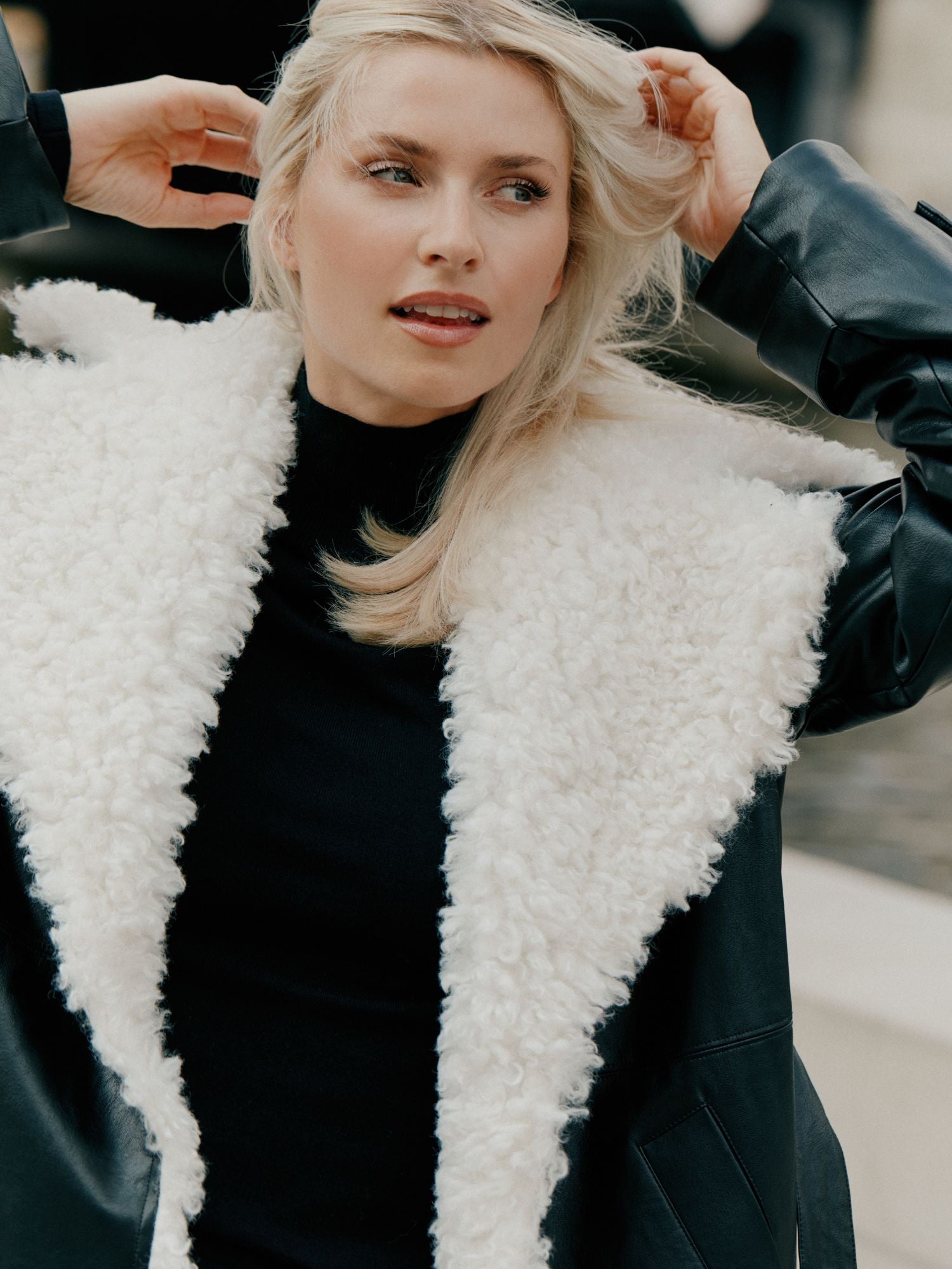 Eine Frau mit langen blonden Haaren in einem schwarzen Oberteil und dem Janique Coat von LeGer by Lena Gercke - einer schwarzen Lederjacke mit einem großen weißen Shearling-Kragen - steht im Freien, schaut zur Seite und hebt ihr Haar.