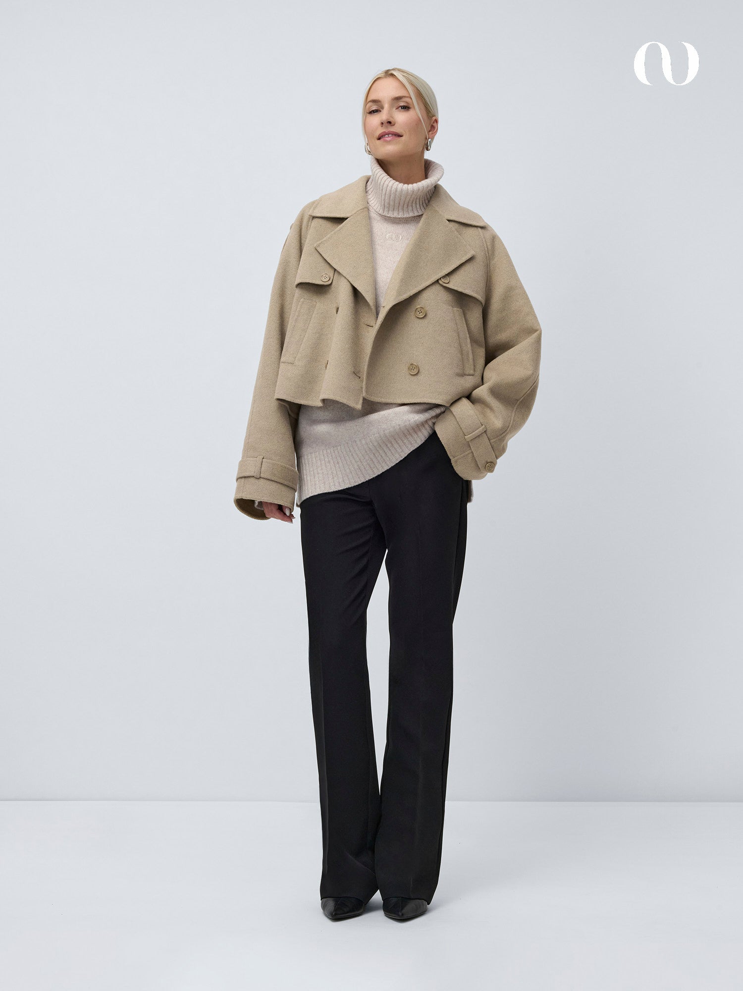 Eine Frau mit zurückgebundenen blonden Haaren lächelt leicht, während sie eine beigefarbene Kurzjacke über dem Erica-Pullover von LeGer by Lena Gercke mit extra langen Ärmeln modelliert. Dazu trägt sie eine schwarze Hose und Schuhe vor einem schlichten hellgrauen Hintergrund.