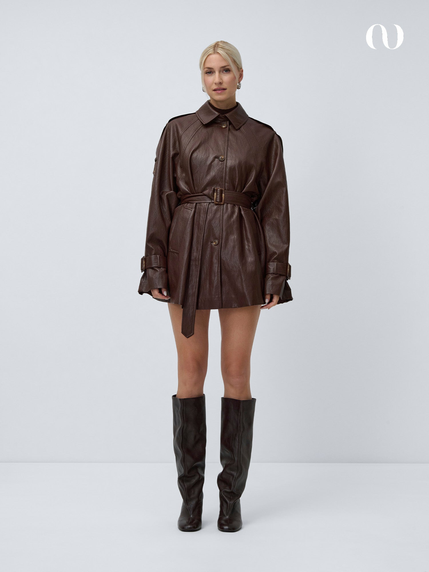 Eine Person steht vor einem schlichten weißen Hintergrund und trägt die dunkelbraune Dinorah Jacke von LeGer by Lena Gercke mit Bindegürtel, langen Ärmeln, Knopfdetails und einer lässigen Passform, gepaart mit passenden kniehohen Stiefeln. Ihr Haar ist zu einer Hochsteckfrisur gestylt.