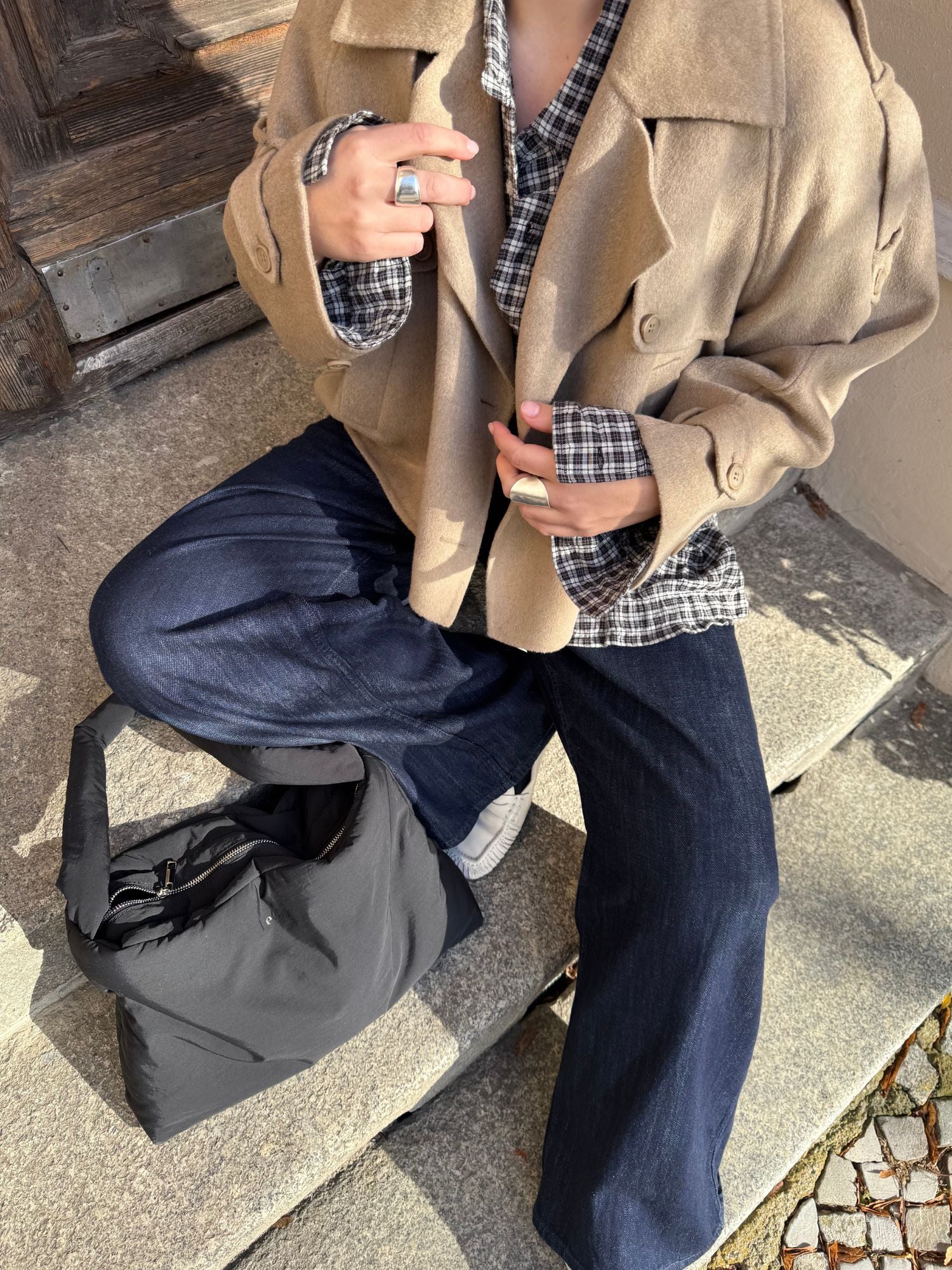 Ein Damenmodel sitzt auf steinernen Stufen und trägt die Elianna Jacket von LeGer by Lena Gercke über einem karierten Hemd, Jeans mit weitem Bein und weißen Sneakers. Silberne Ringe schimmern im Sonnenlicht, eine schwarze Tasche liegt neben ihr.