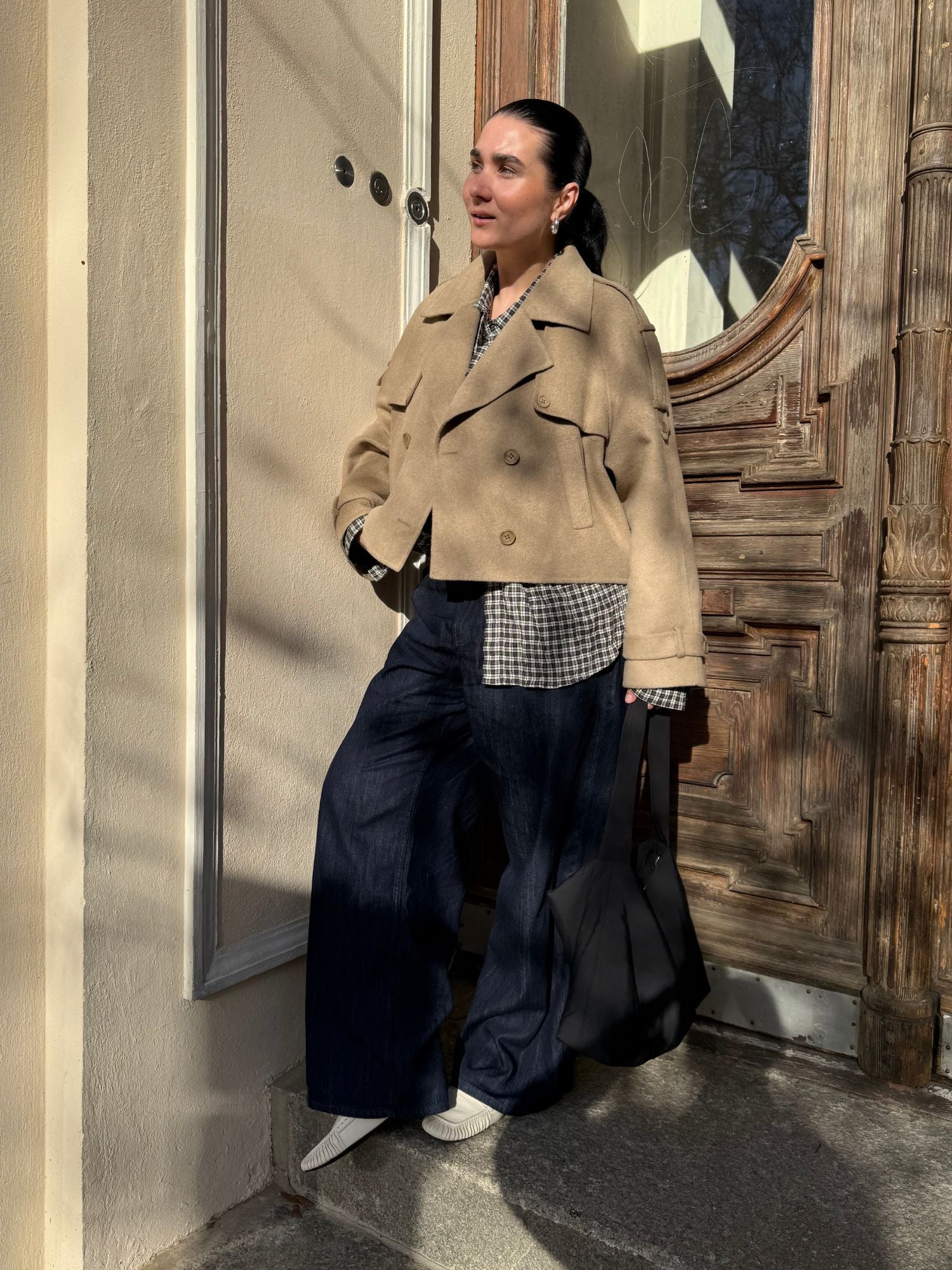 Eine Frau steht im Freien im Sonnenlicht vor einer geschnitzten Holztür. Sie trägt eine beigefarbene Elianna Jacket von LeGer by Lena Gercke über einem karierten Hemd, dunkle Jeans mit weitem Bein, weiße Schuhe und eine schwarze Tragetasche.