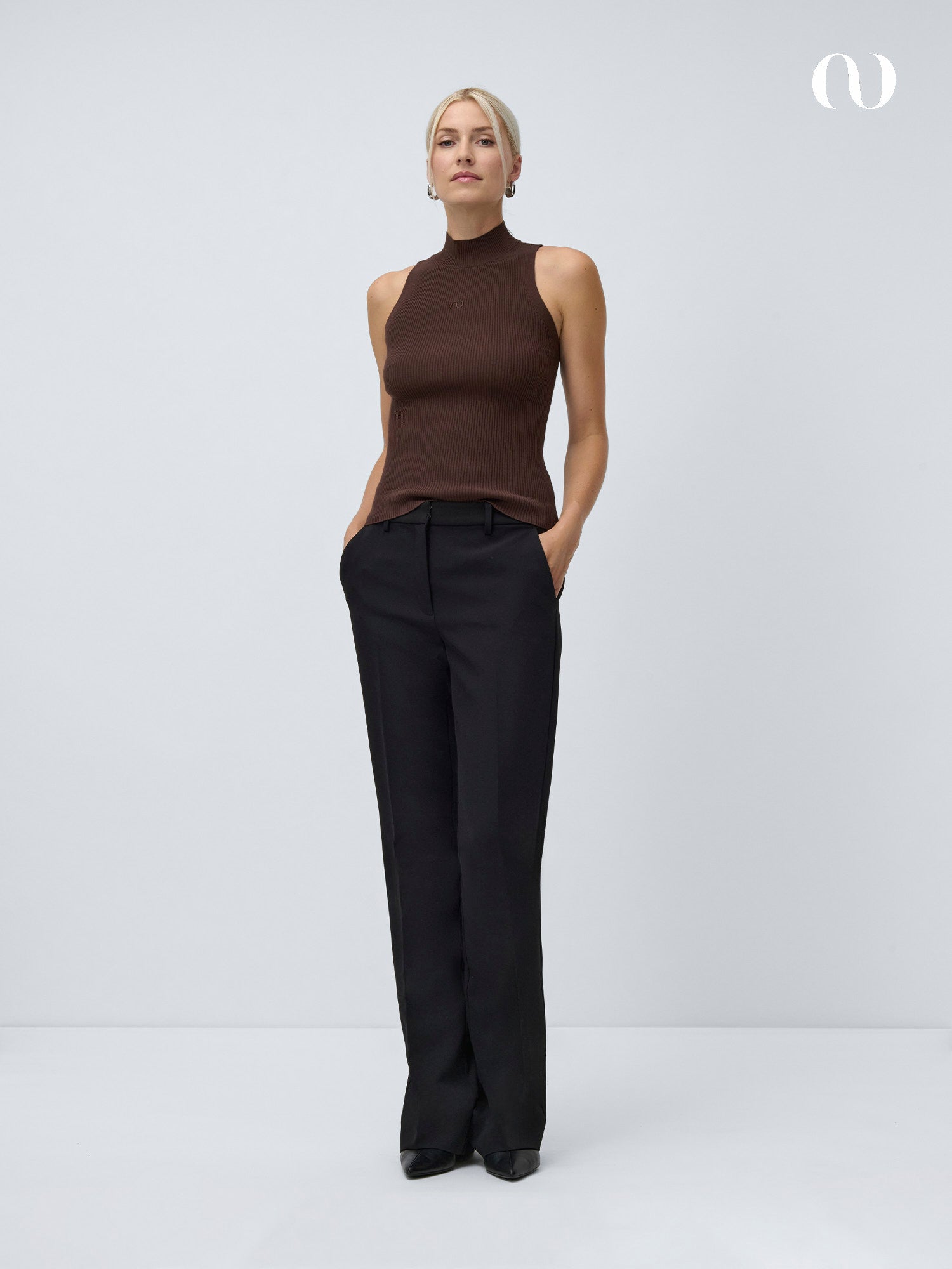Eine Frau mit zurückgebundenen blonden Haaren steht vor einem weißen Hintergrund und trägt einen ärmellosen braunen Rollkragenpullover und die Fatou Pants Tall von LeGer by Lena Gercke in Schwarz. Sie hat die Hände in den Taschen und einen neutralen Gesichtsausdruck.
