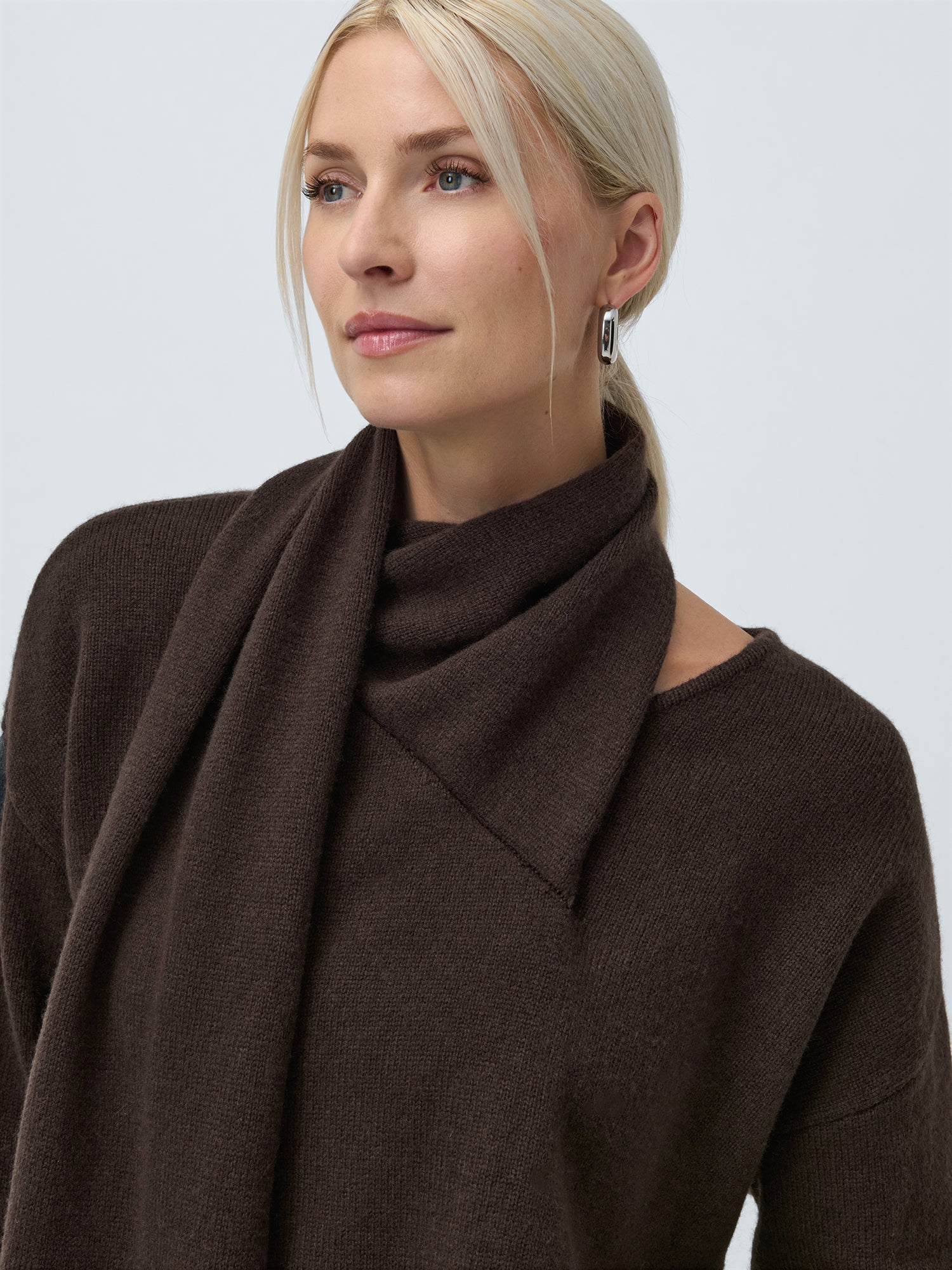 Eine Frau mit blondem Haar in einem niedrigen Pferdeschwanz trägt den locker sitzenden, dunkelbraunen Helga-Pullover von LeGer by Lena Gercke und einen passenden Schal und blickt sanft zur Seite vor einem schlichten hellen Hintergrund.