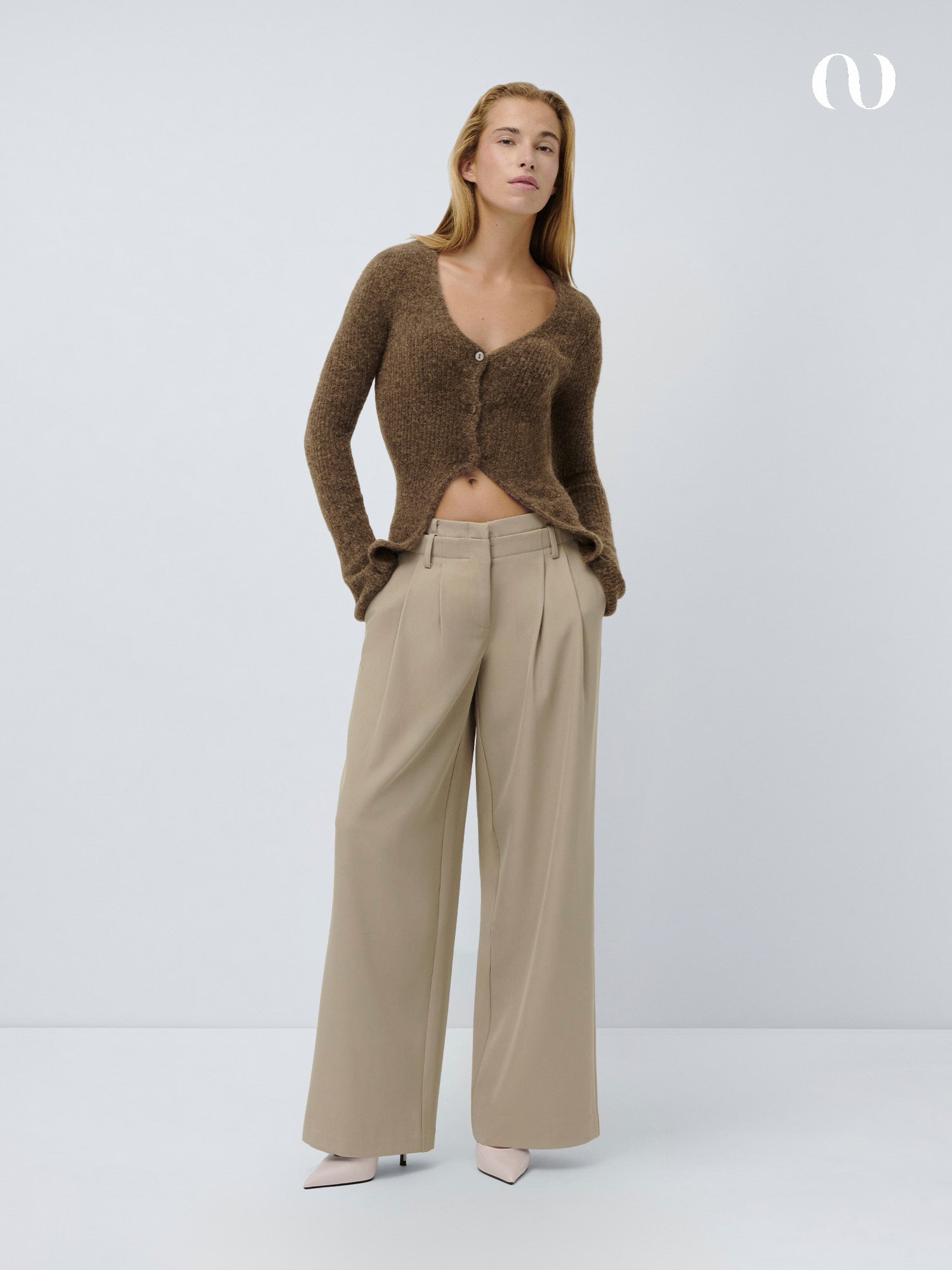 Eine Frau steht vor einem schlichten hellen Hintergrund und trägt eine braune Strickjacke mit tiefem V-Ausschnitt und die Marcelina-Hose von LeGer by Lena Gercke. Sie vervollständigt den Look mit hellen, spitz zulaufenden Absätzen.