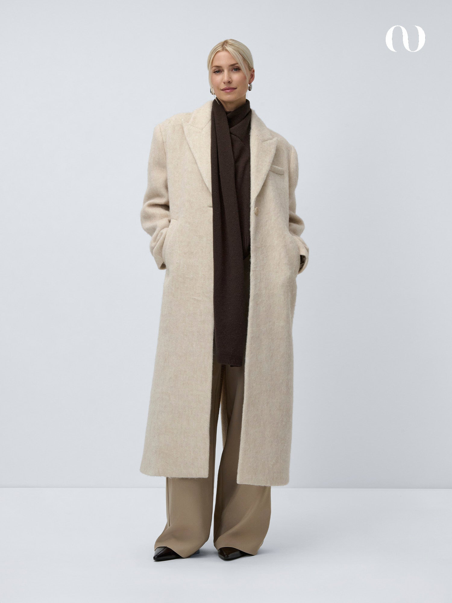 Eine Person steht vor einem schlichten, hellen Hintergrund und trägt den Linda-Mantel von LeGer by Lena Gercke über einem dunkelbraunen Oberteil und einem Schal, gepaart mit einer weiten, beigefarbenen Hose und schwarzen spitzen Schuhen. Die Hände in den Taschen, das Haar für eine elegante Silhouette nach hinten gezogen.