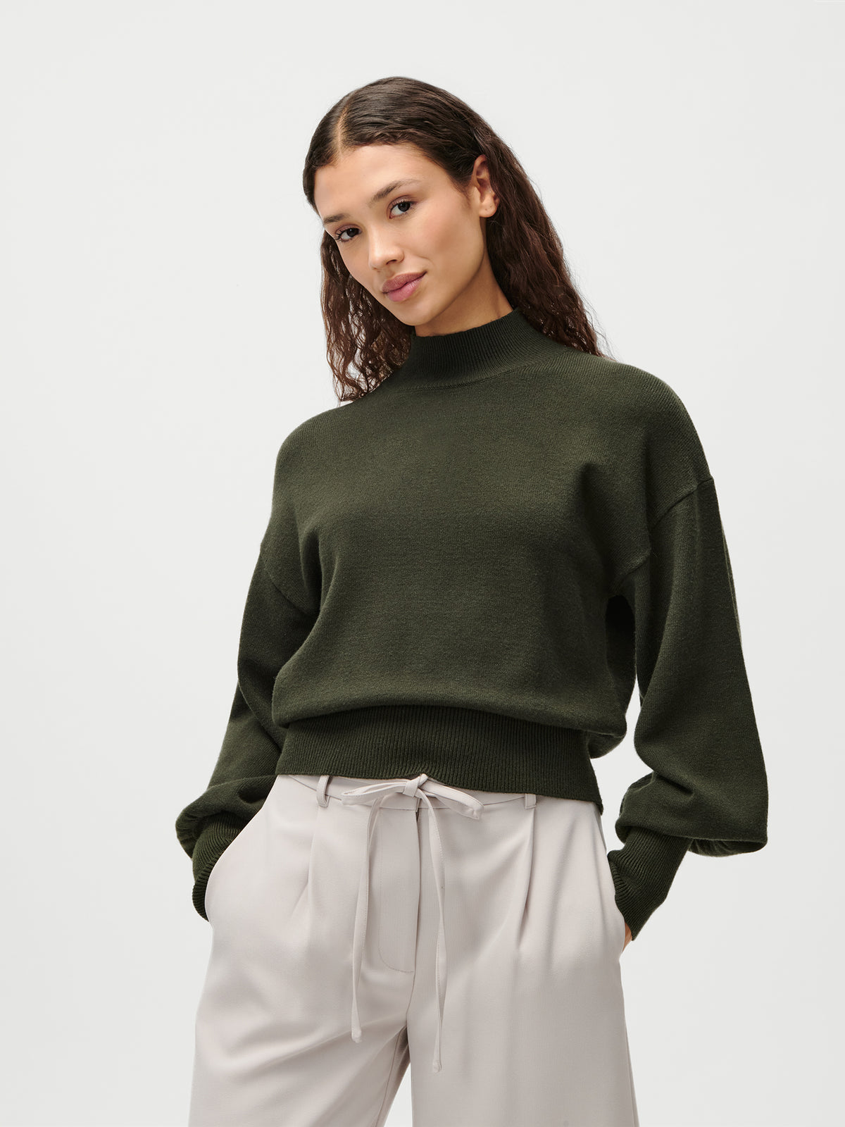 Eine Frau mit langen, welligen braunen Haaren trägt den Pullover „Penelope“ von LeGer by Lena Gercke in Olivgrün mit einem stilvollen Stehkragen, dazu eine hellbeige Hose mit hoher Taille und Bindegürtel; sie steht vor einem schlichten weißen Hintergrund.