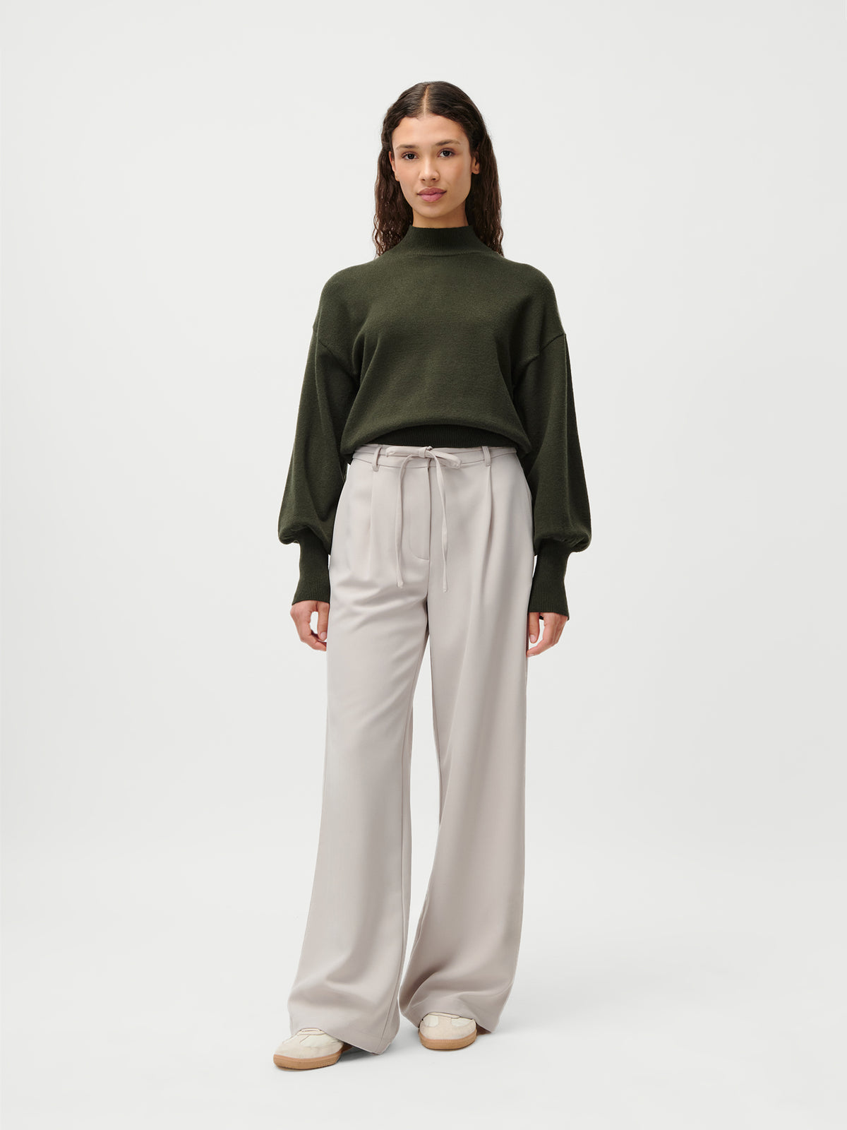 Vor einem schlichten weißen Hintergrund steht eine Frau mit dem Blick nach vorne. Sie trägt den Pullover „Penelope“ von LeGer by Lena Gercke in Olivgrün mit Ballonärmeln und Stehkragen, dazu eine hellgraue Hose mit weitem Bein und weiße Sneaker.