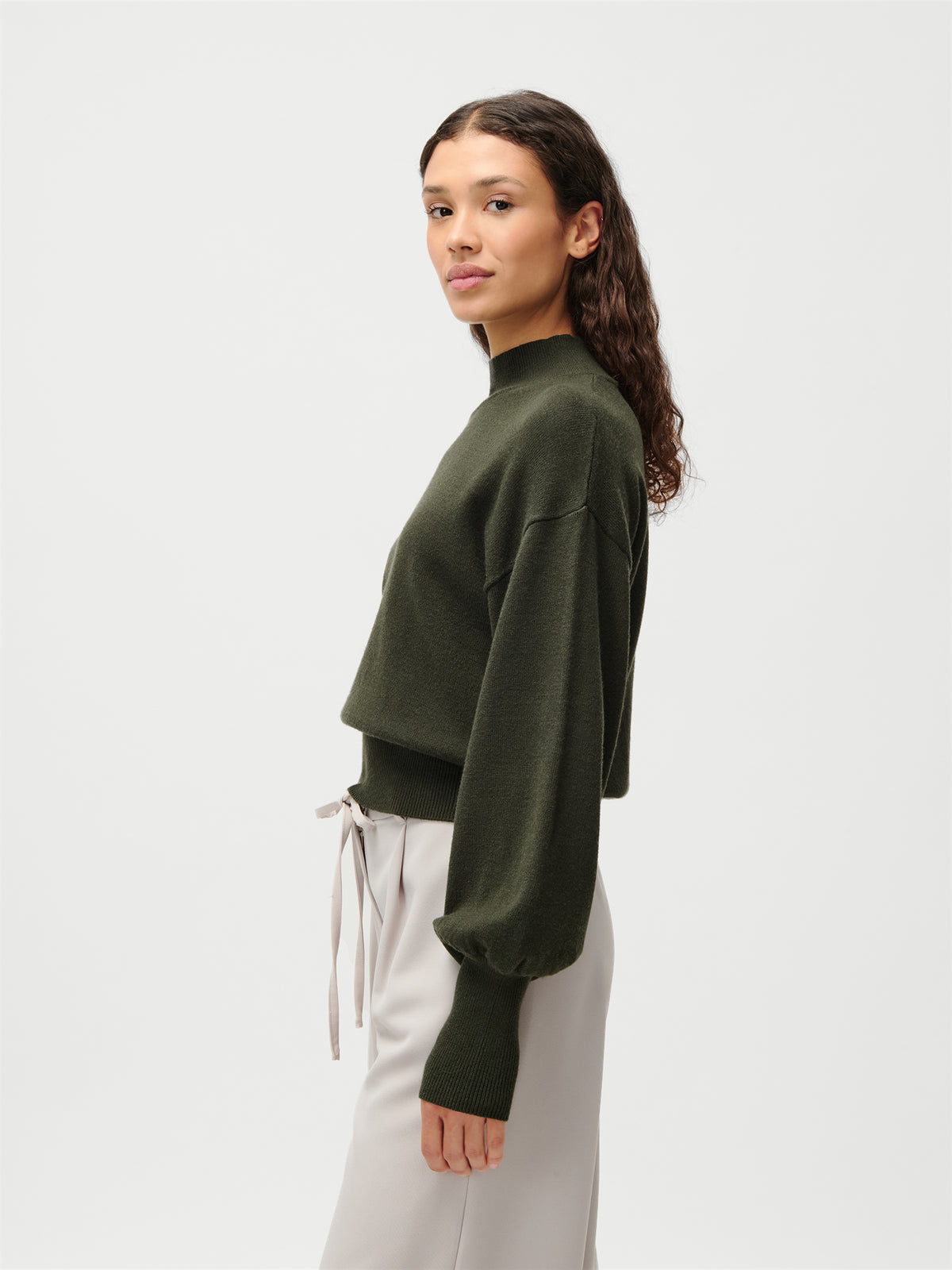 Eine Person mit langen, welligen braunen Haaren trägt den Pullover „Penelope“ aus der Kollektion „LeGer by Lena Gercke“ in Olivgrün mit Puffärmeln, dazu eine helle Hose. Sie steht seitlich vor einem schlichten weißen Hintergrund.