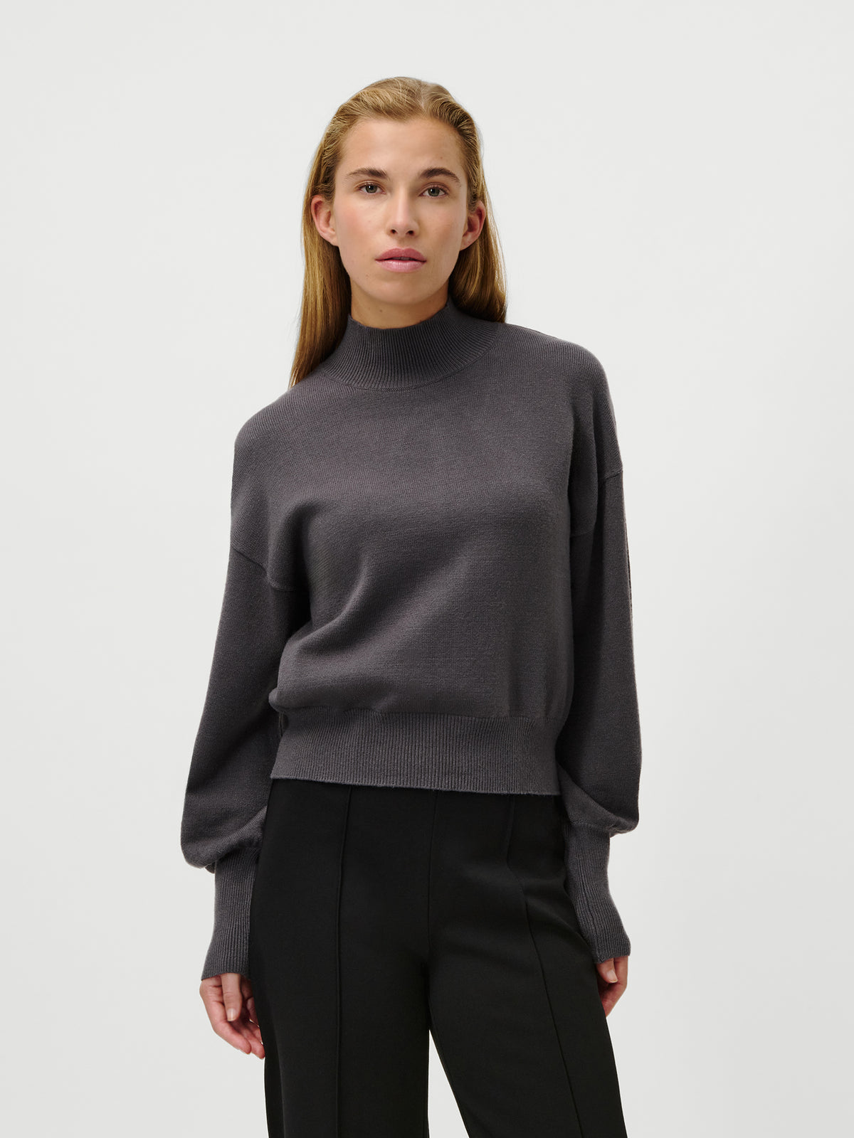 Eine Person mit langen, glatten Haaren trägt den Pullover „Penelope“ von LeGer by Lena Gercke in Dunkelgrau mit langen Ärmeln und Stehkragen, dazu eine schwarze Hose, vor einem schlichten weißen Hintergrund.
