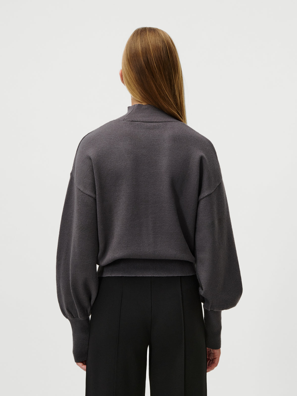 Eine Person mit glattem, langem, hellbraunem Haar steht mit dem Rücken zur Kamera und trägt den Pullover „Penelope“ von LeGer by Lena Gercke – einen locker sitzenden, dunkelgrauen, kurz geschnittenen Pullover mit Stehkragen –, dazu eine schwarze Hose vor einem schlichten, hellen Hintergrund.