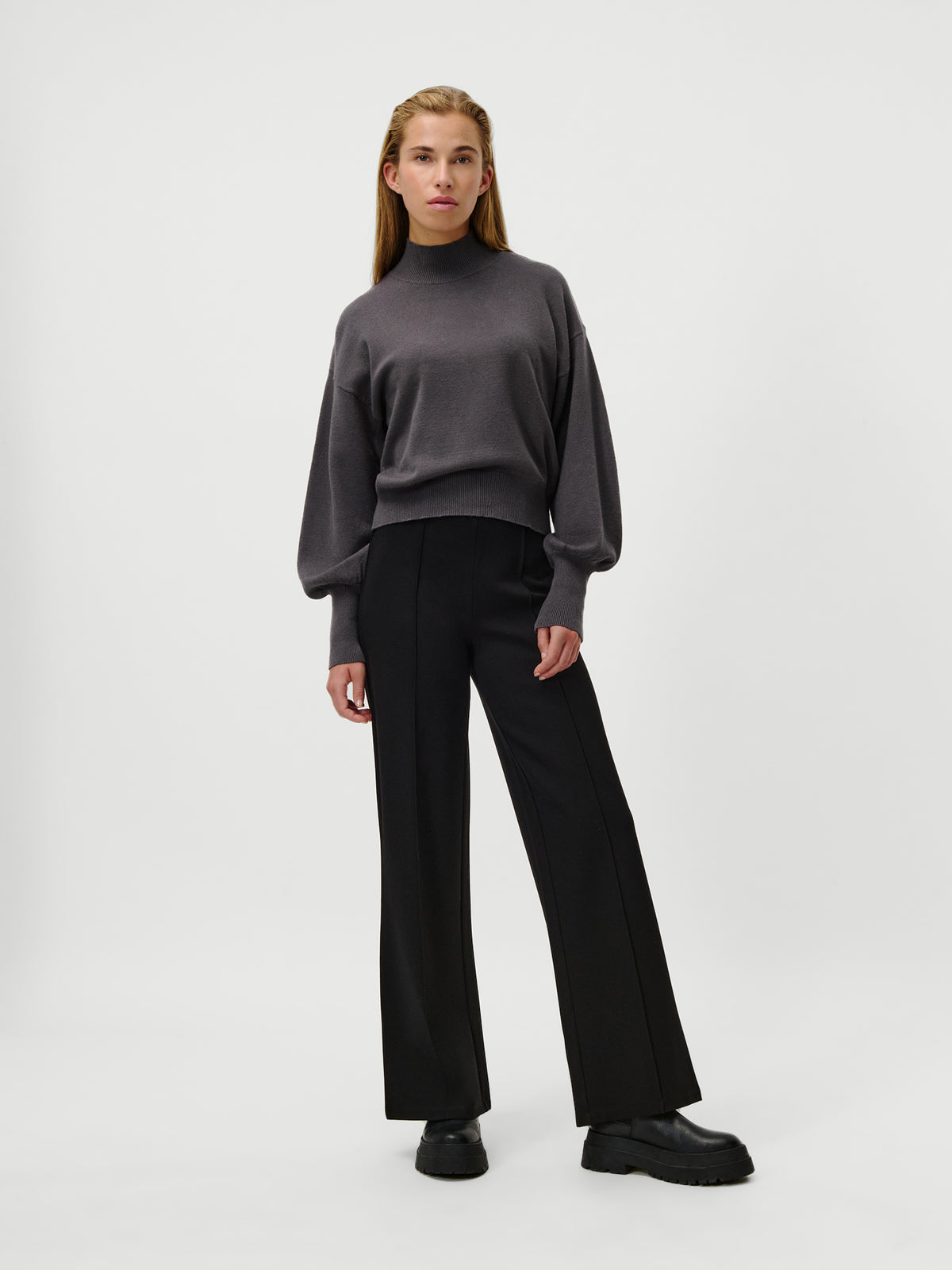 Eine Frau steht vor einem schlichten weißen Hintergrund und trägt den Pullover „Penelope“ von LeGer by Lena Gercke – einen kurzen Pullover mit Stehkragen in Dunkelgrau –, kombiniert mit einer schwarzen Hose mit weitem Bein und schwarzen klobigen Schuhen.