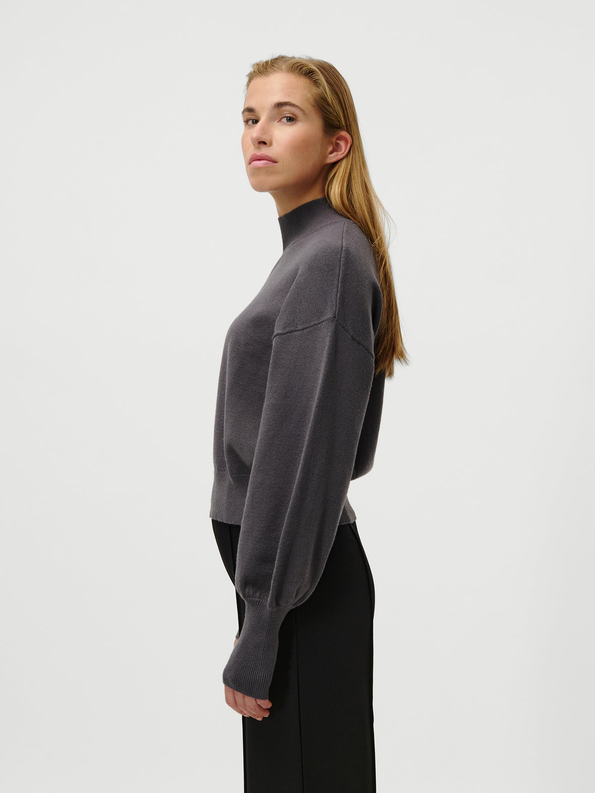Eine Person mit langen, glatten Haaren steht im Profil vor einem schlichten Hintergrund und trägt den Pullover „Penelope“ von LeGer by Lena Gercke – einen dunkelgrauen, kurzen Pullover mit Stehkragen – sowie eine schwarze Hose.