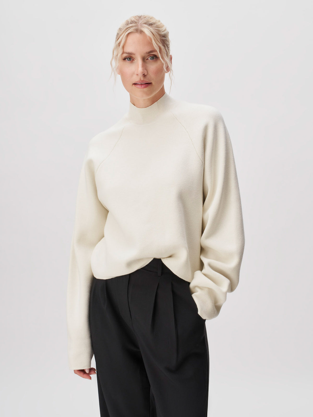 Eine Person steht vor einem hellen Hintergrund und trägt den Pullover „Heather“ von LeGer by Lena Gercke, einen cremefarbenen Pullover mit langen Ärmeln und normaler Länge, der sich durch einen hohen Ausschnitt und eine lockere Passform auszeichnet. Der Look wird mit einer locker sitzenden schwarzen Hose kombiniert, und die Person wirkt entspannt, während sie eine Hand in der Hosentasche hat.